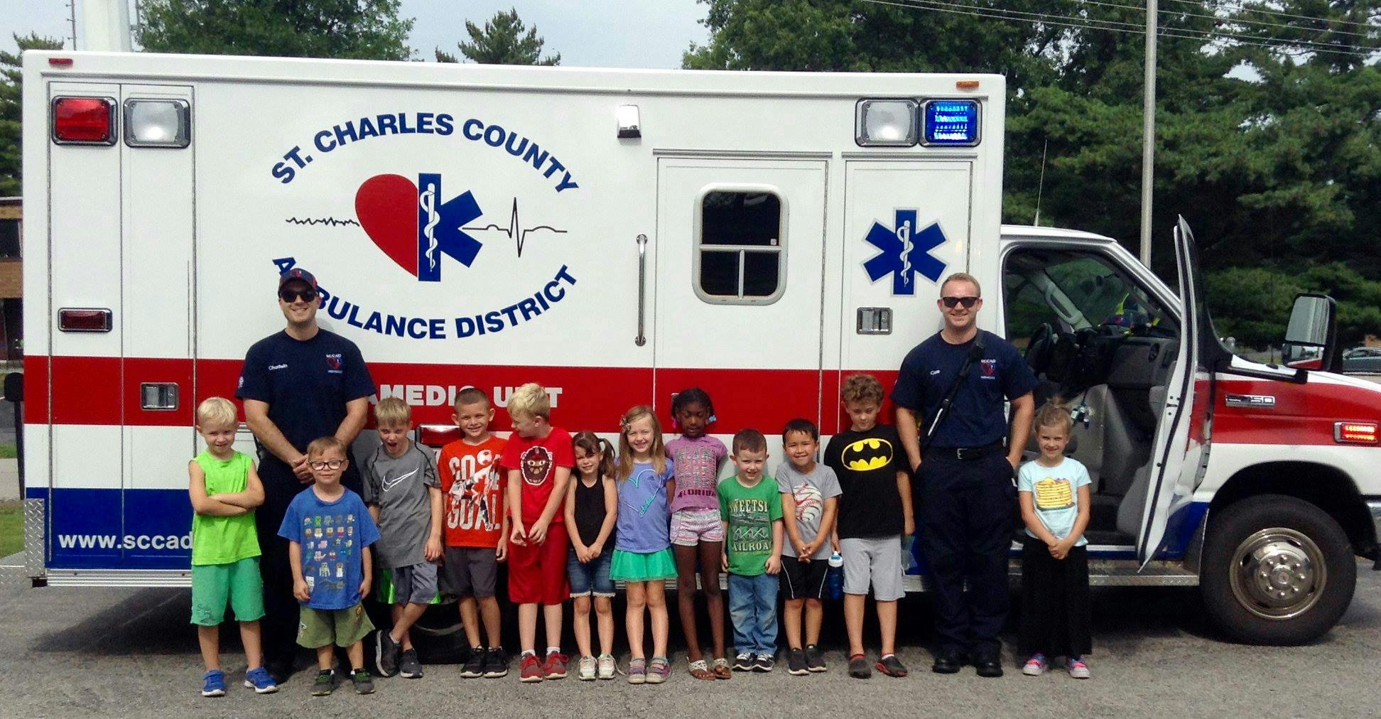 Two people and multiple children standing in front of an ambulance