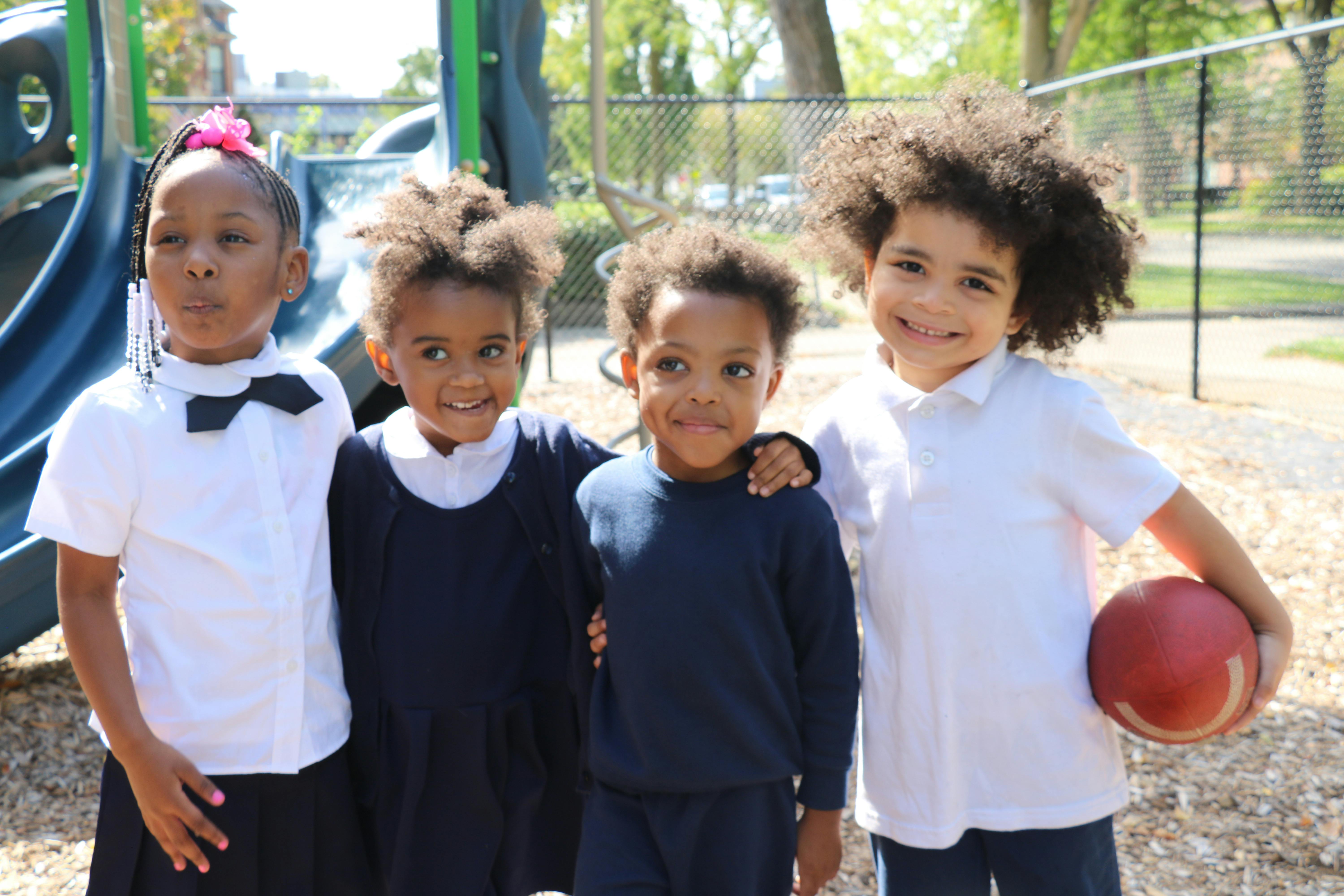 Four children standing together on a playground, one holding a red ball