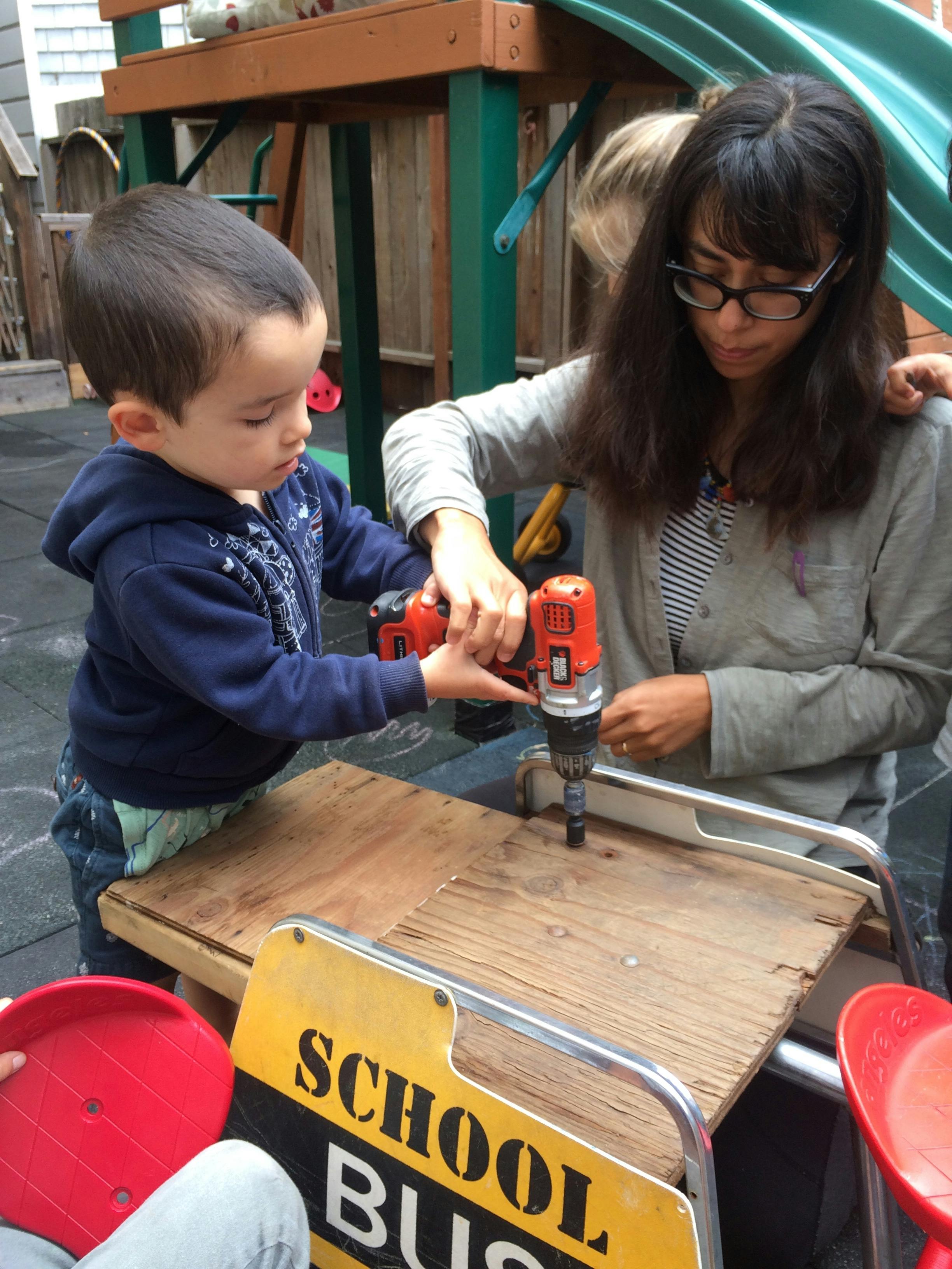Adult and child using a drill on a piece of wood, with a school bus sign below