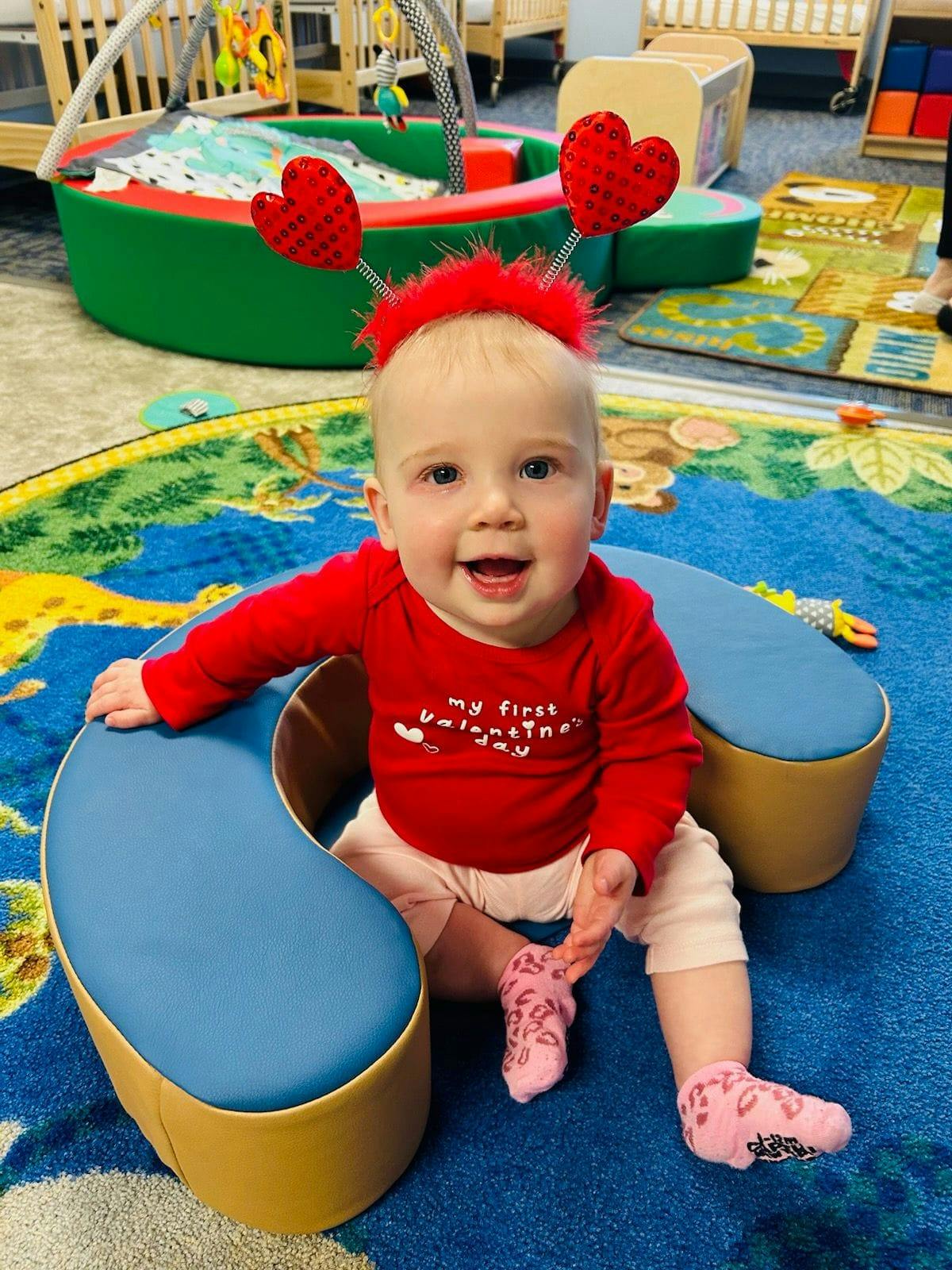 Child wearing a red shirt and heart headband sitting on a blue mat