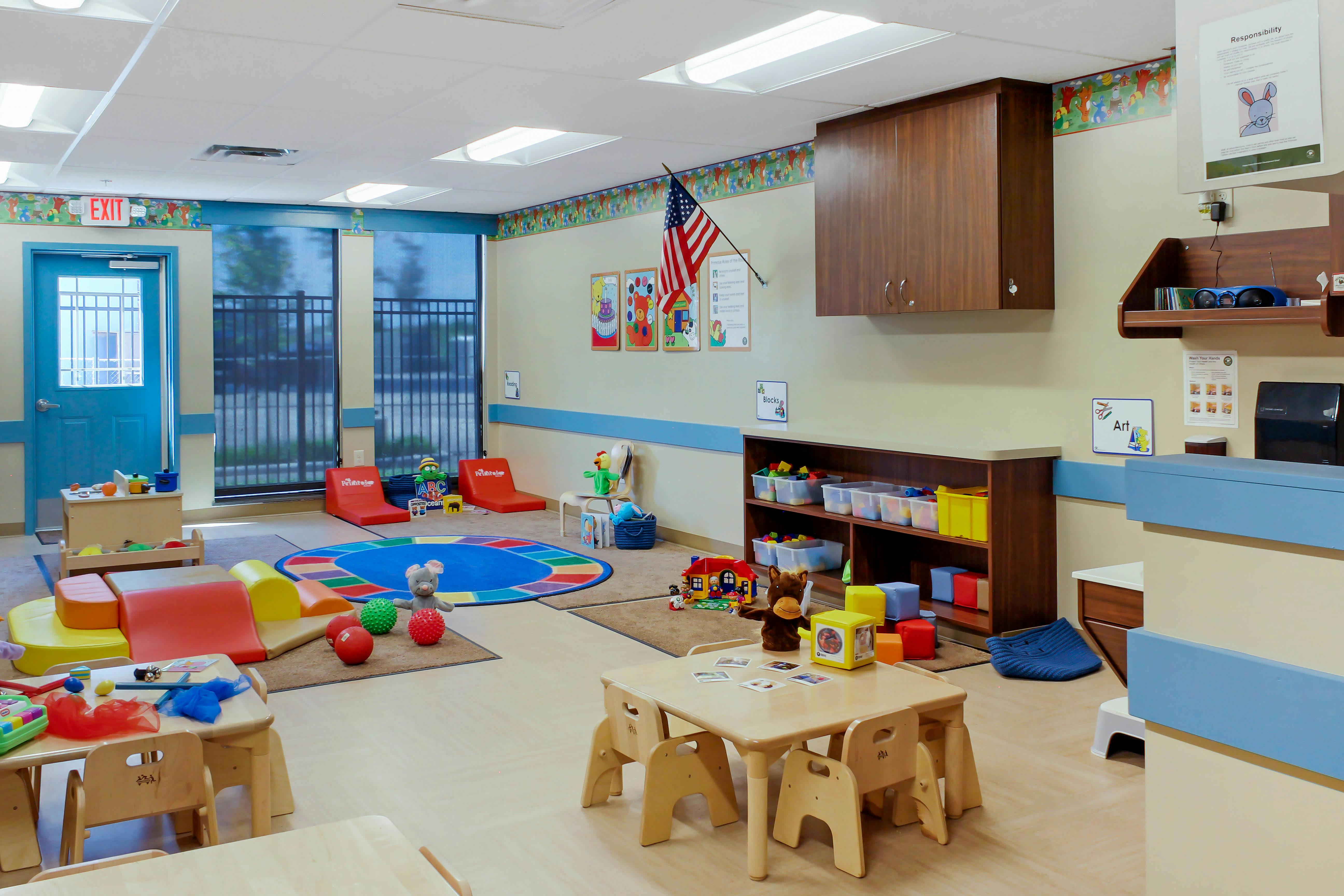 Childcare classroom with small tables and chairs, colorful play mats, and toys