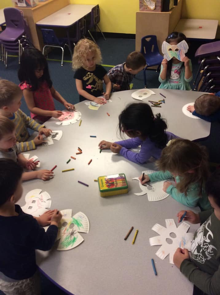 Children sitting around a table coloring paper masks with crayons