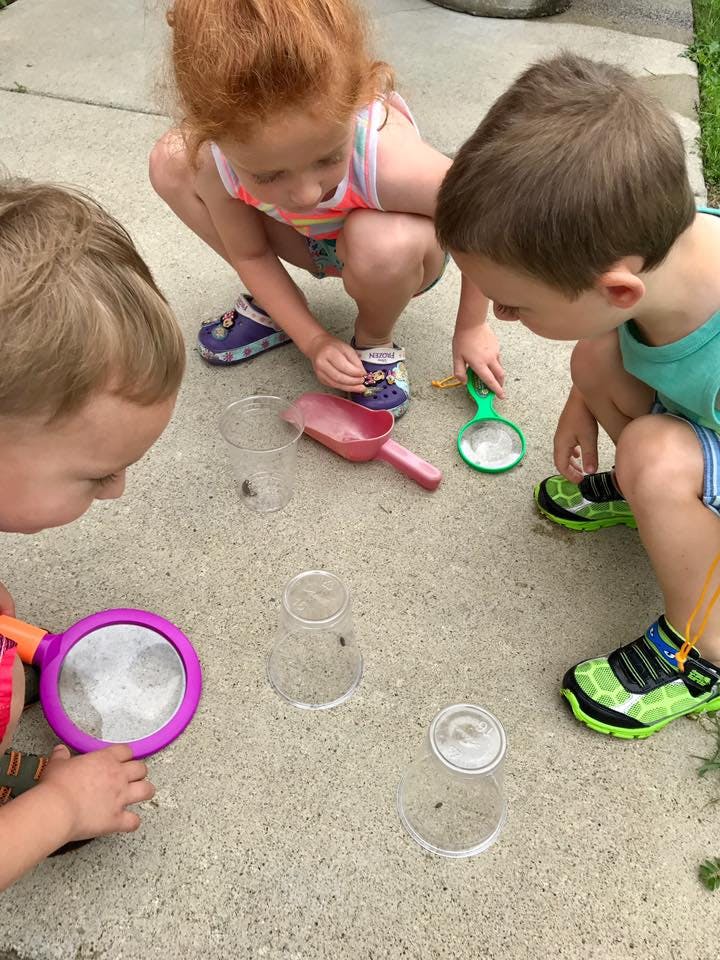 Three children looking at insects on the ground with magnifying glasses