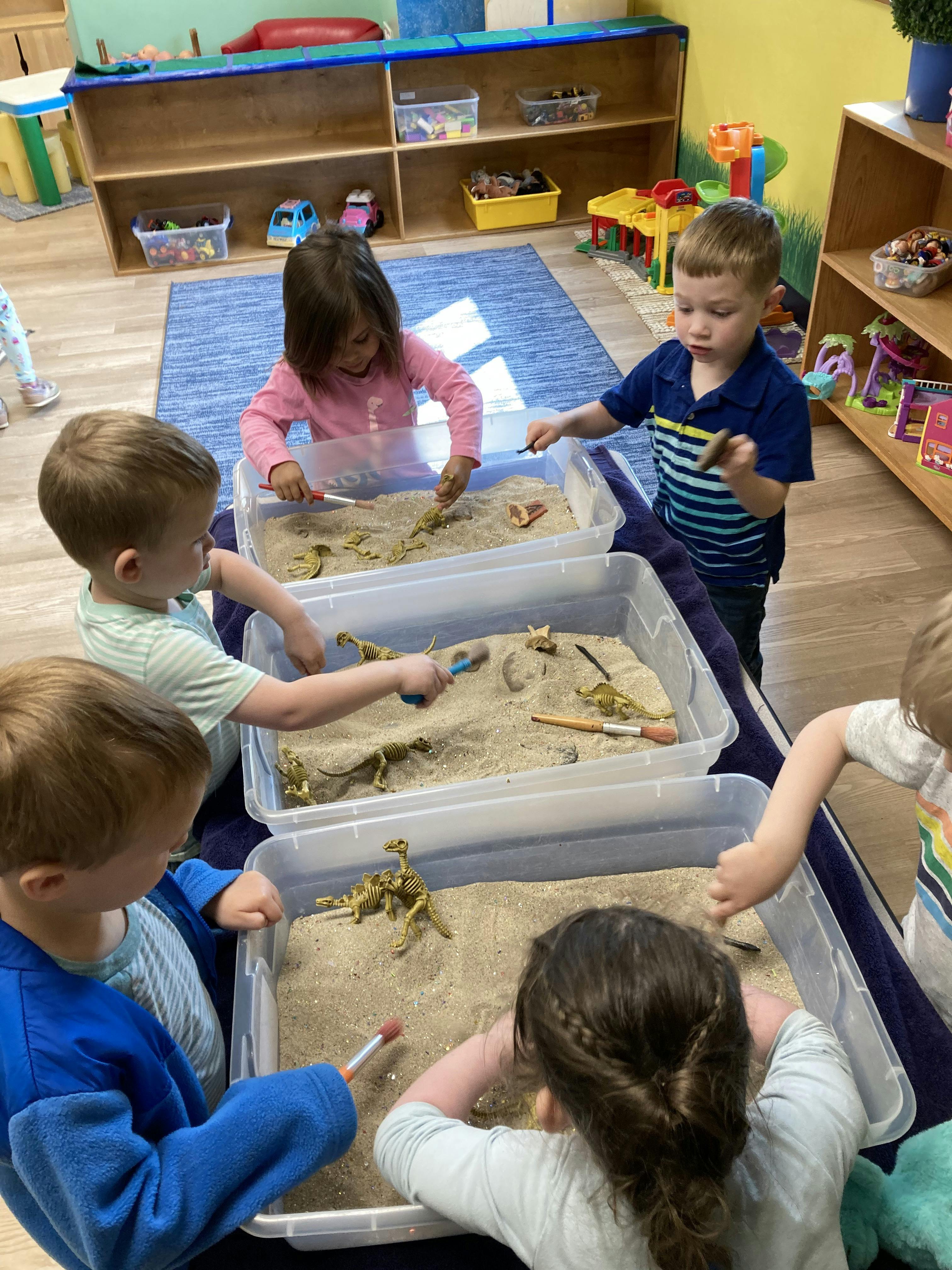 Children playing with sand and toy dinosaurs in plastic bins