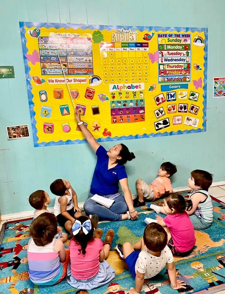Person pointing at a calendar on a wall with children sitting on the floor