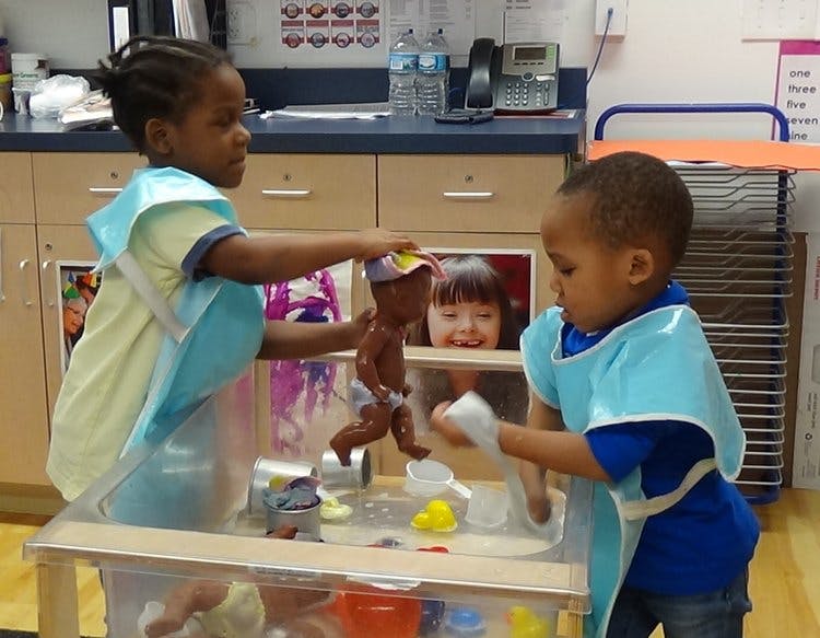 Two children playing with dolls and water at a table