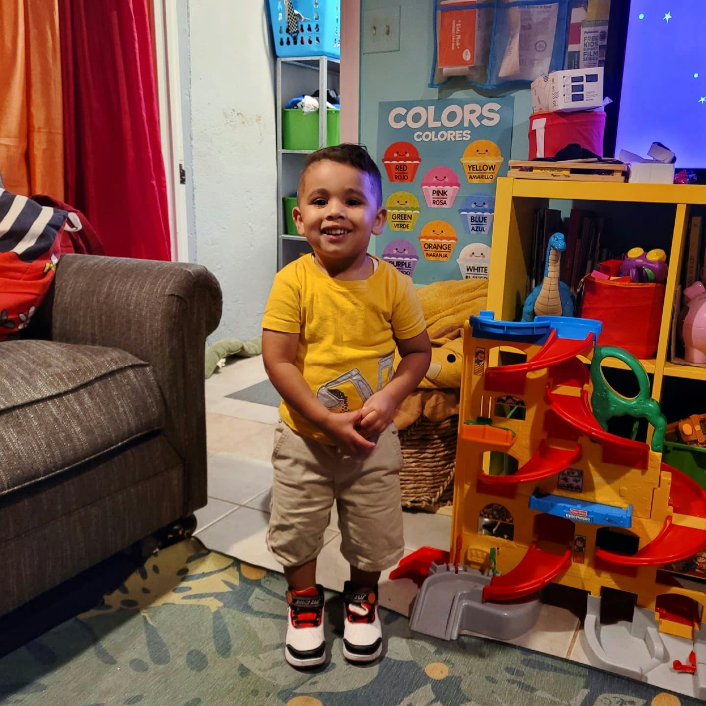 Child standing in a room with a toy car ramp and a "COLORS" poster