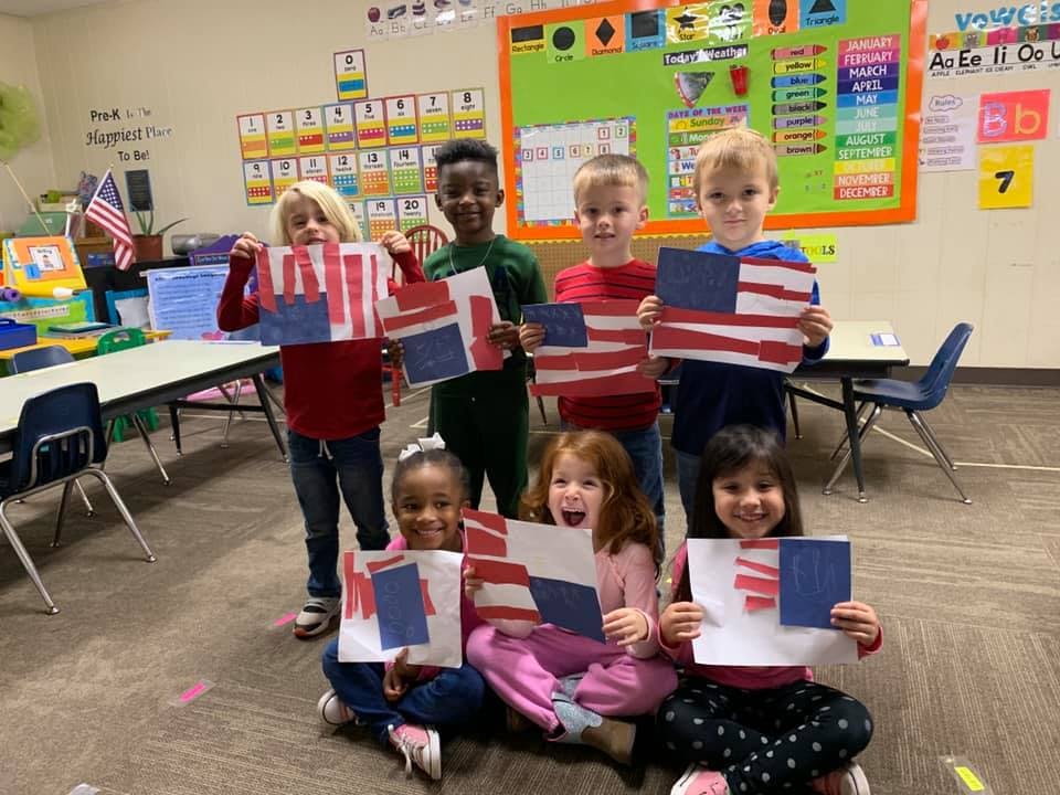 Six children and one teen holding up American flag crafts in a classroom