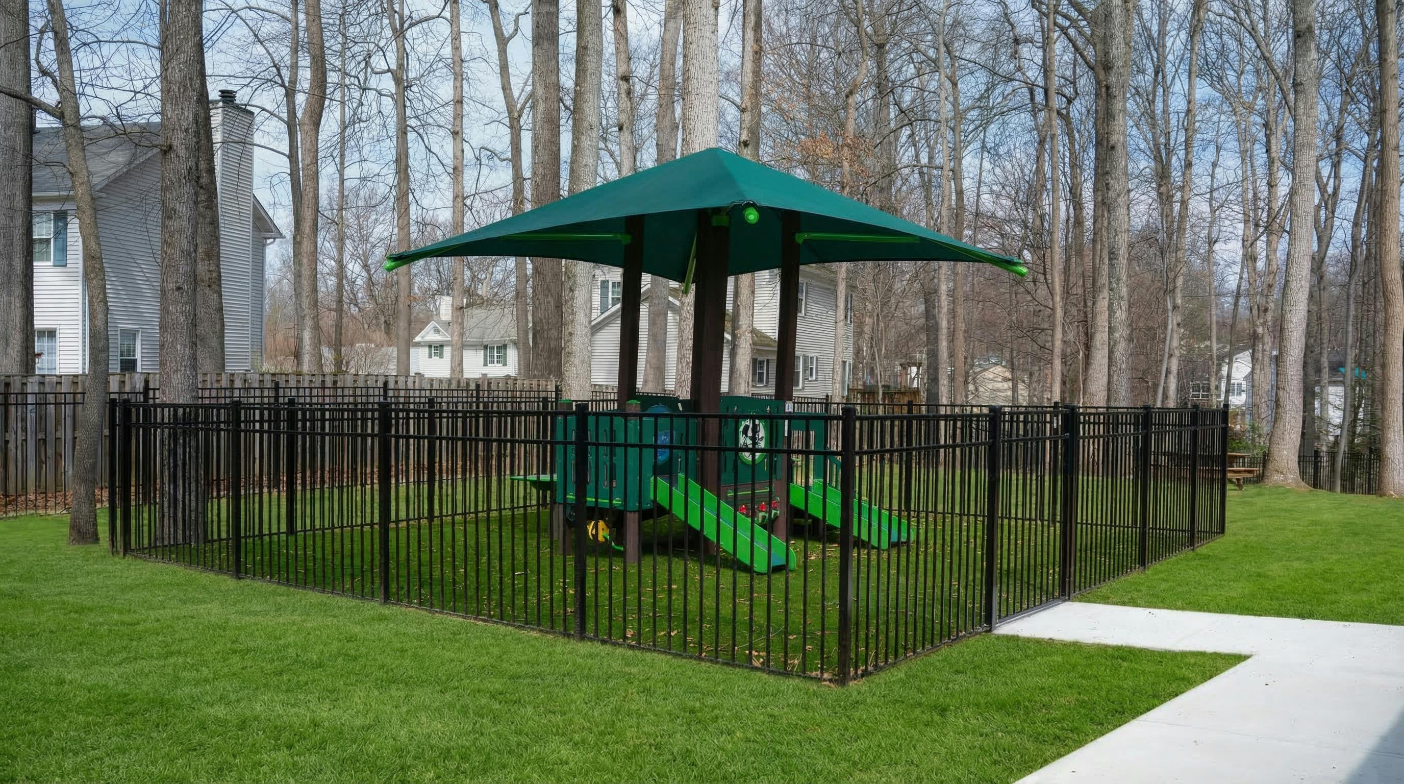 A fenced-in green playground with a shade cover and slides in a grassy area