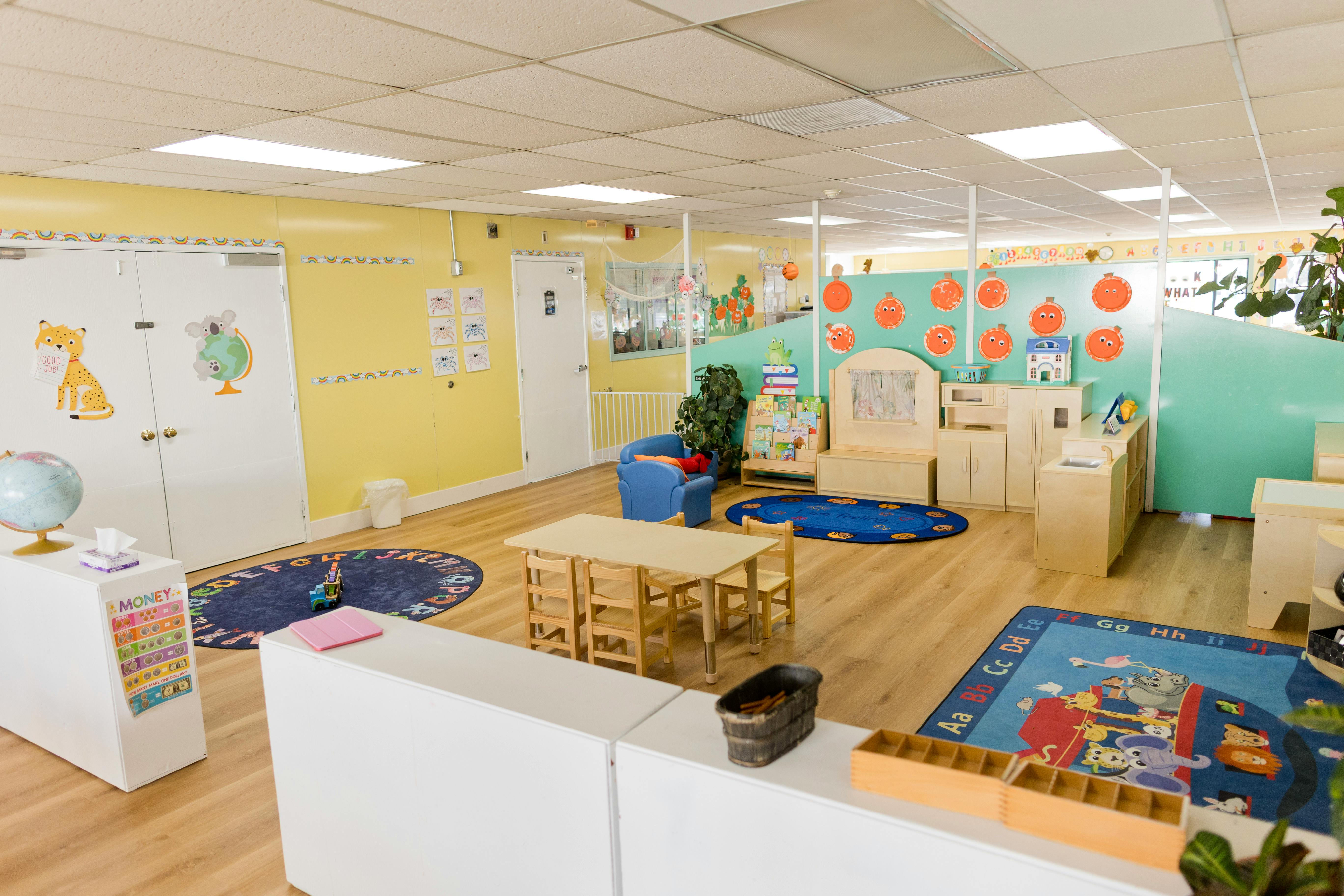 Brightly lit children's classroom with small tables, chairs, and play areas