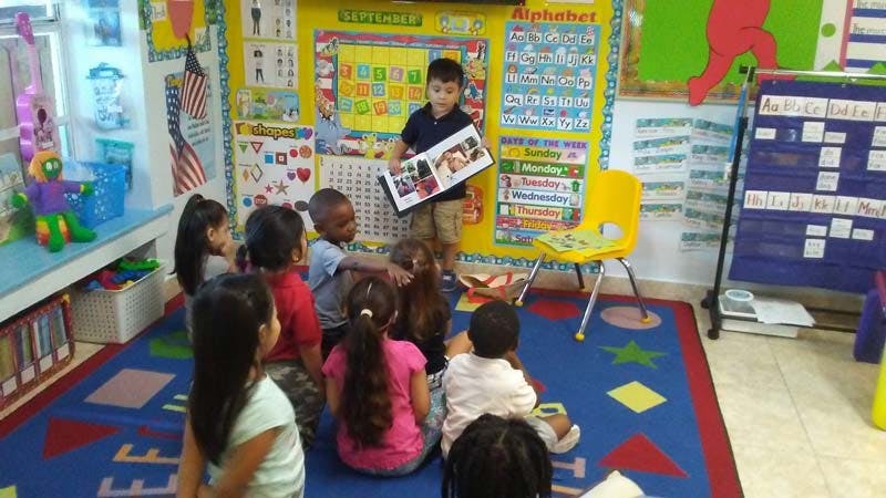 Child holding a book and speaking to other children sitting on a rug