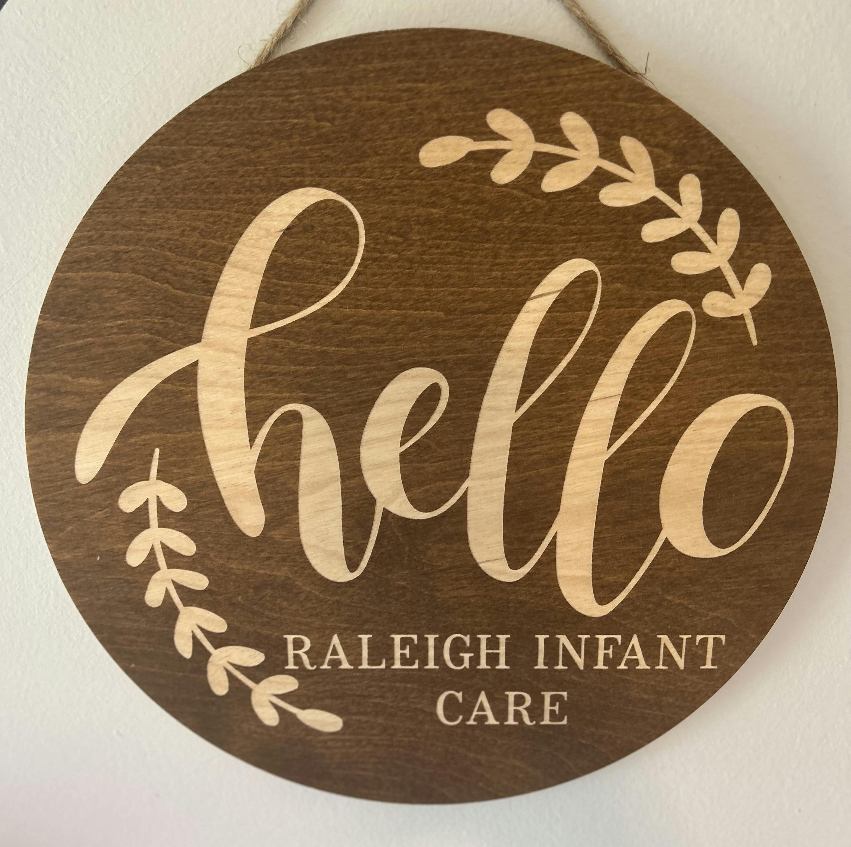 Round wooden sign with the text hello Raleigh Infant Care and leafy accents