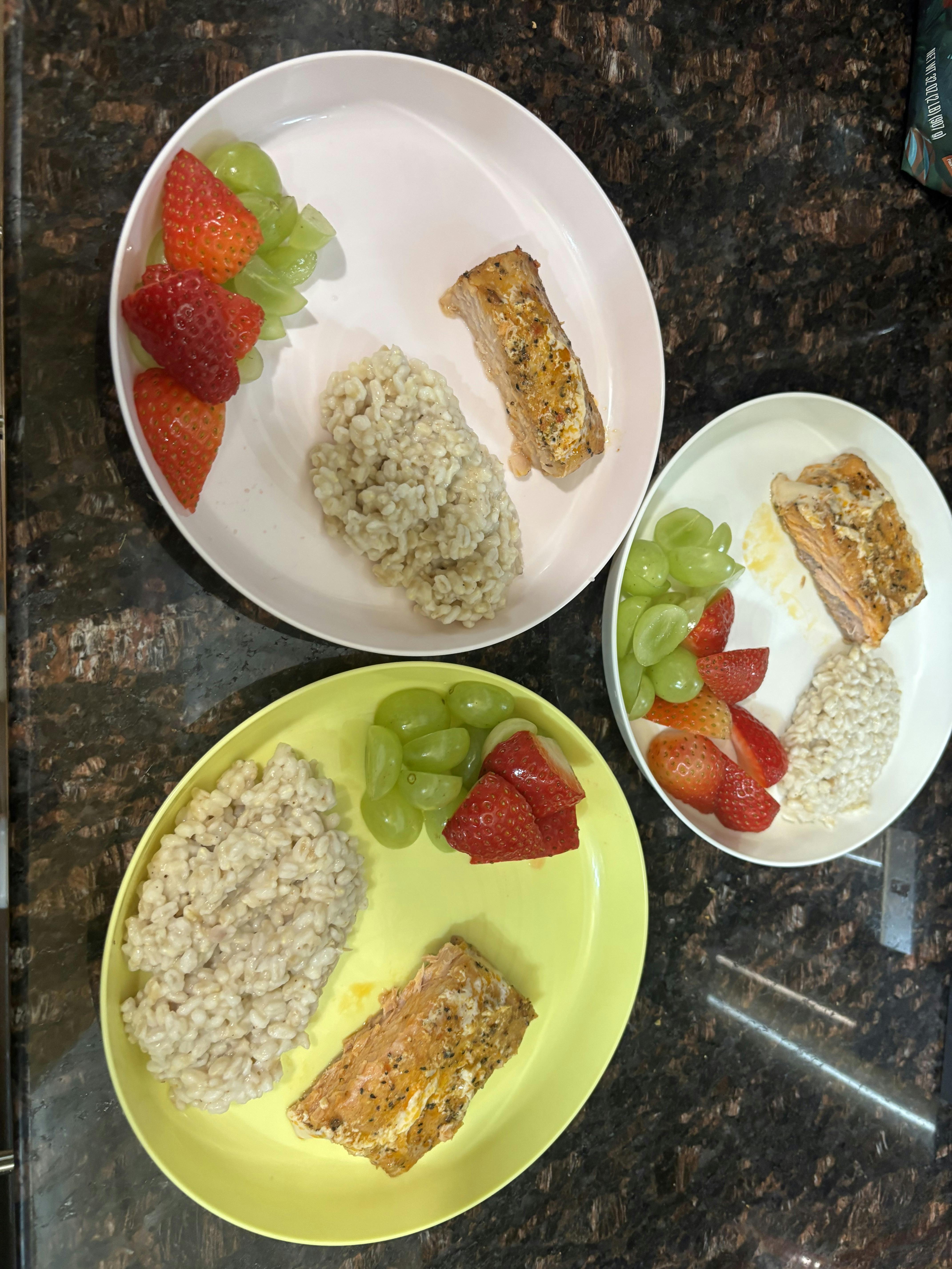 Three plates of salmon, grains, strawberries, and grapes on a kitchen counter