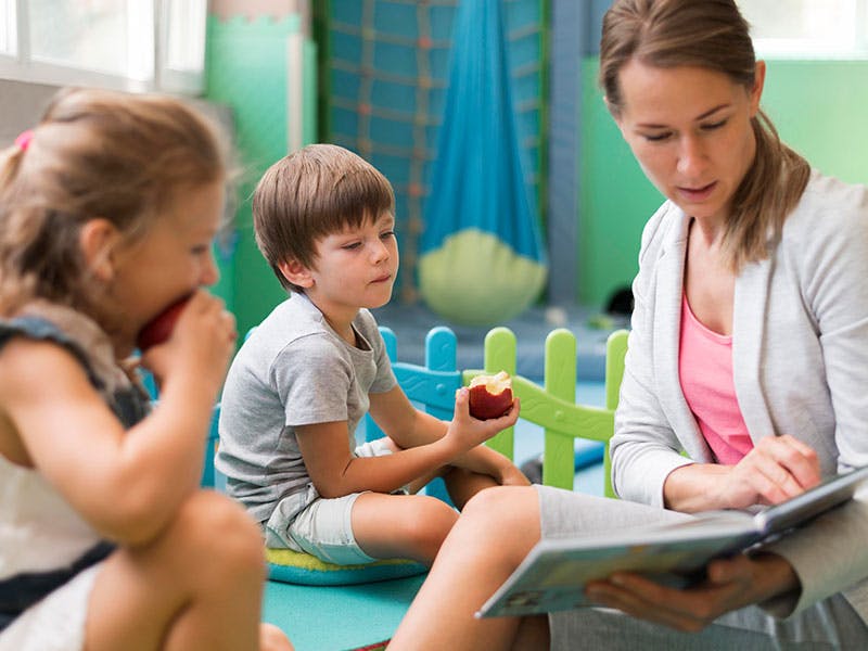 Adult reading a book to two children, one holding an apple