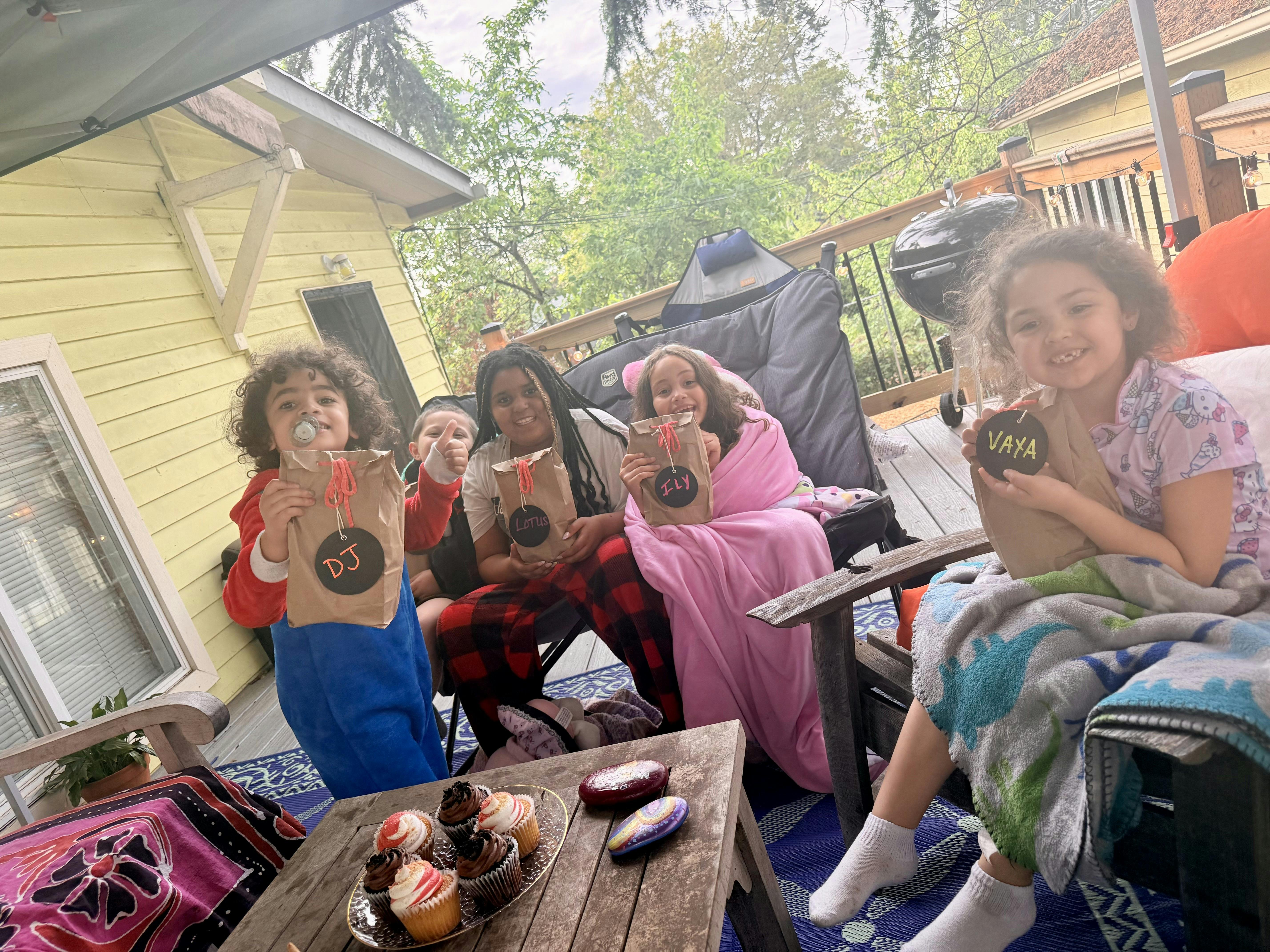 Children sitting on a deck holding personalized brown bags, with cupcakes on a table