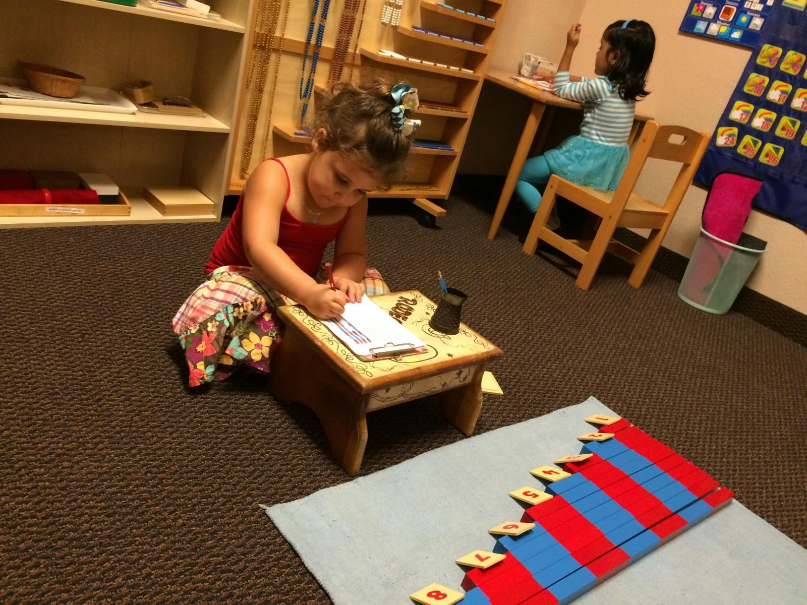 Child writing at a small table on the floor in a classroom