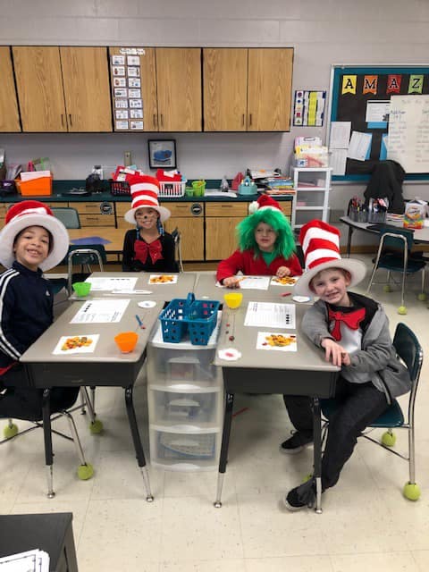 Four children in costume hats sitting at desks in a classroom