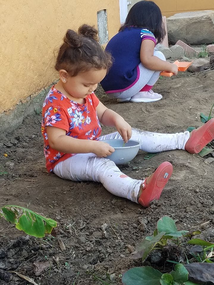 Child sitting on dirt, holding a bowl, with another child in the background