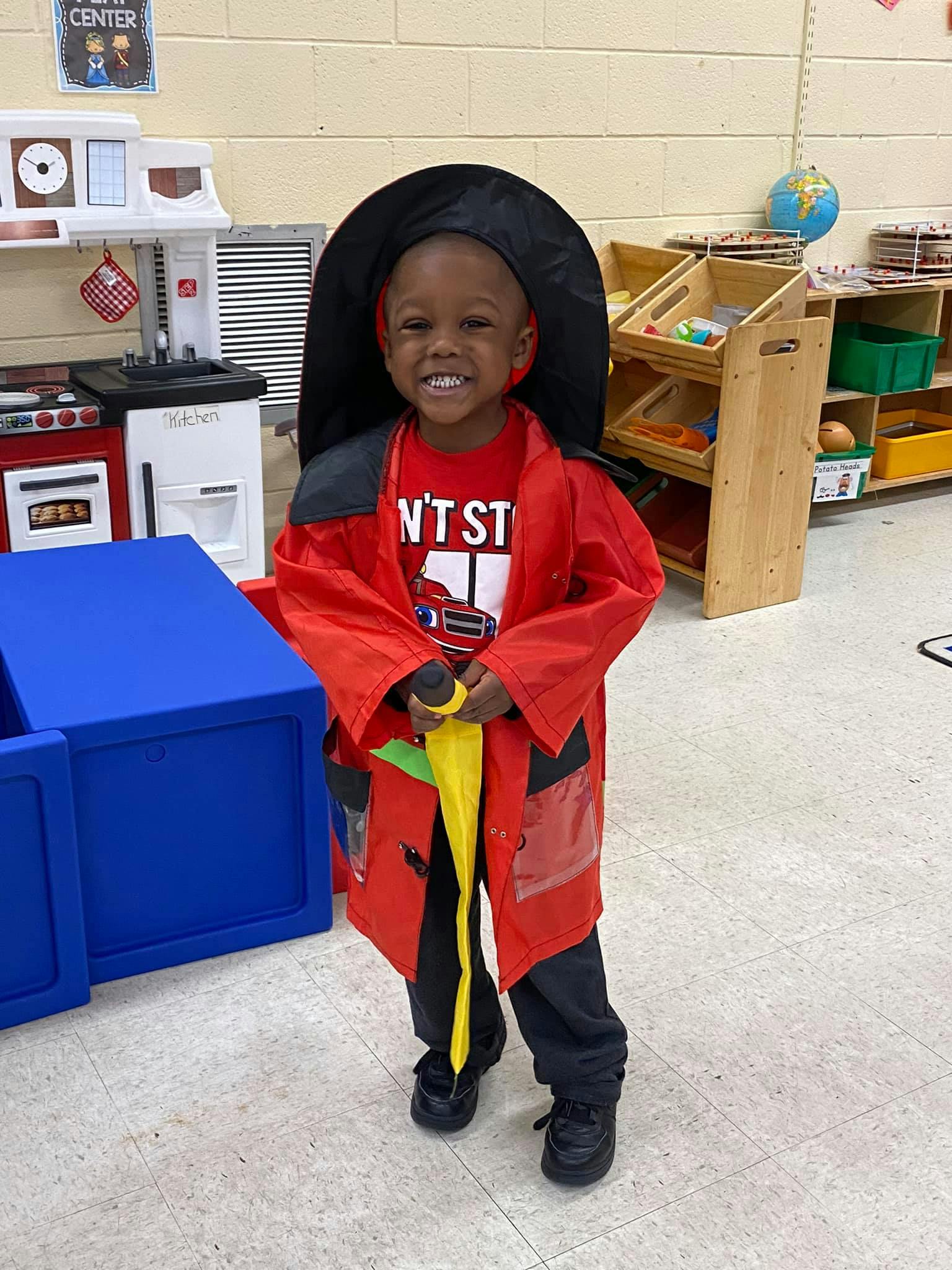 Child in a red firefighter costume holding a yellow hose in a classroom