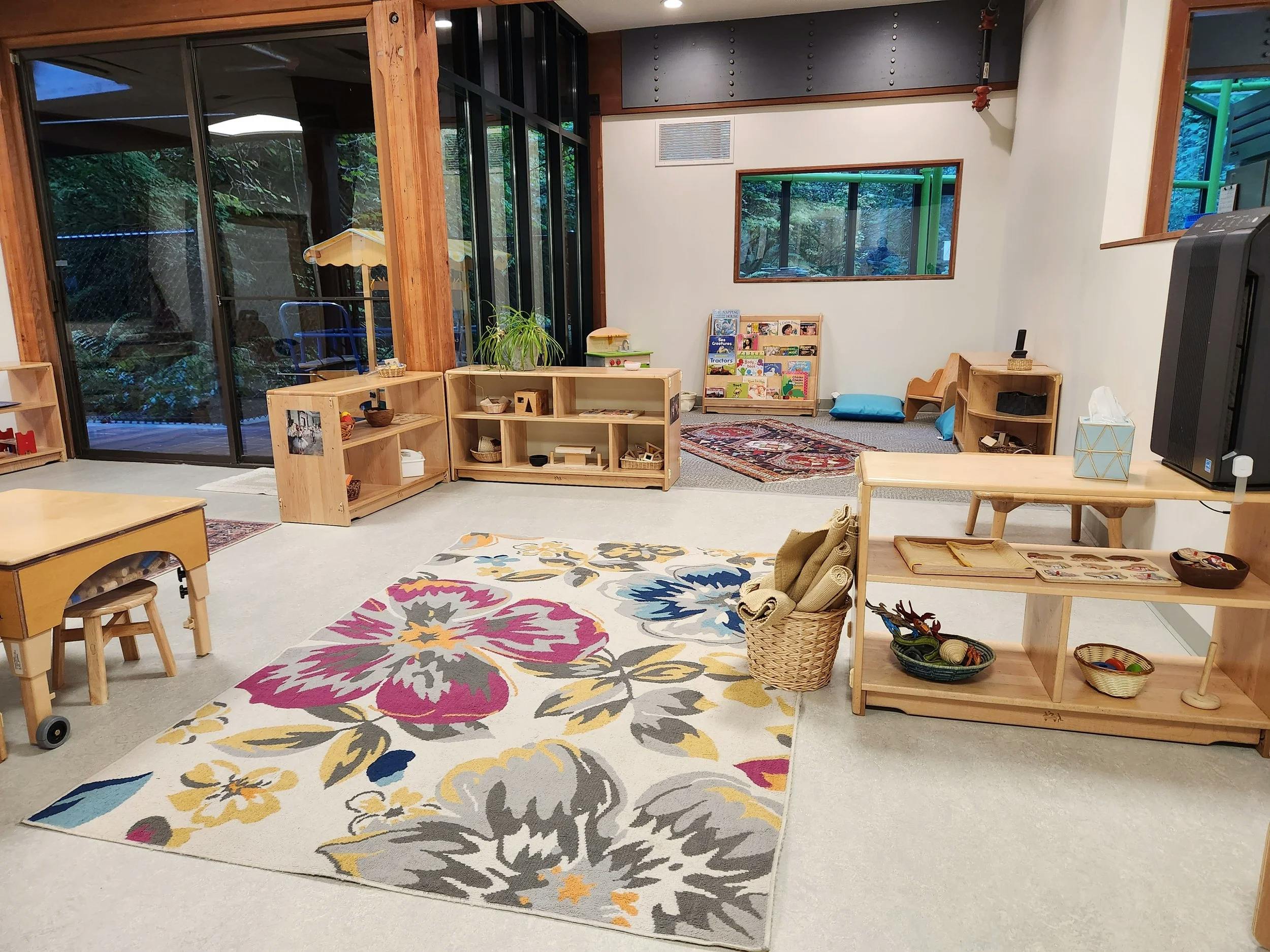 Classroom with wooden shelves, tables, colorful rug, and large windows