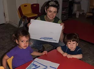 Adult holding up a drawing with two children sitting at a table