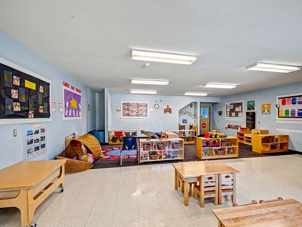 Empty classroom with small tables and chairs, shelves with toys, and posters on the walls