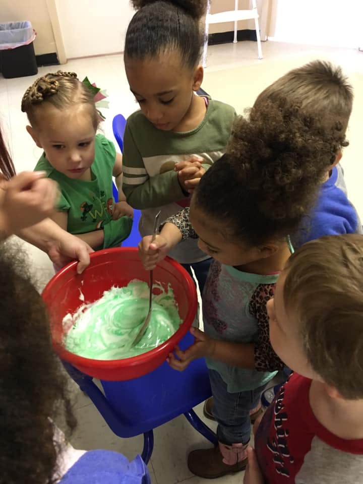 Children gathered around a red bowl, one child stirring green mixture with a spoon