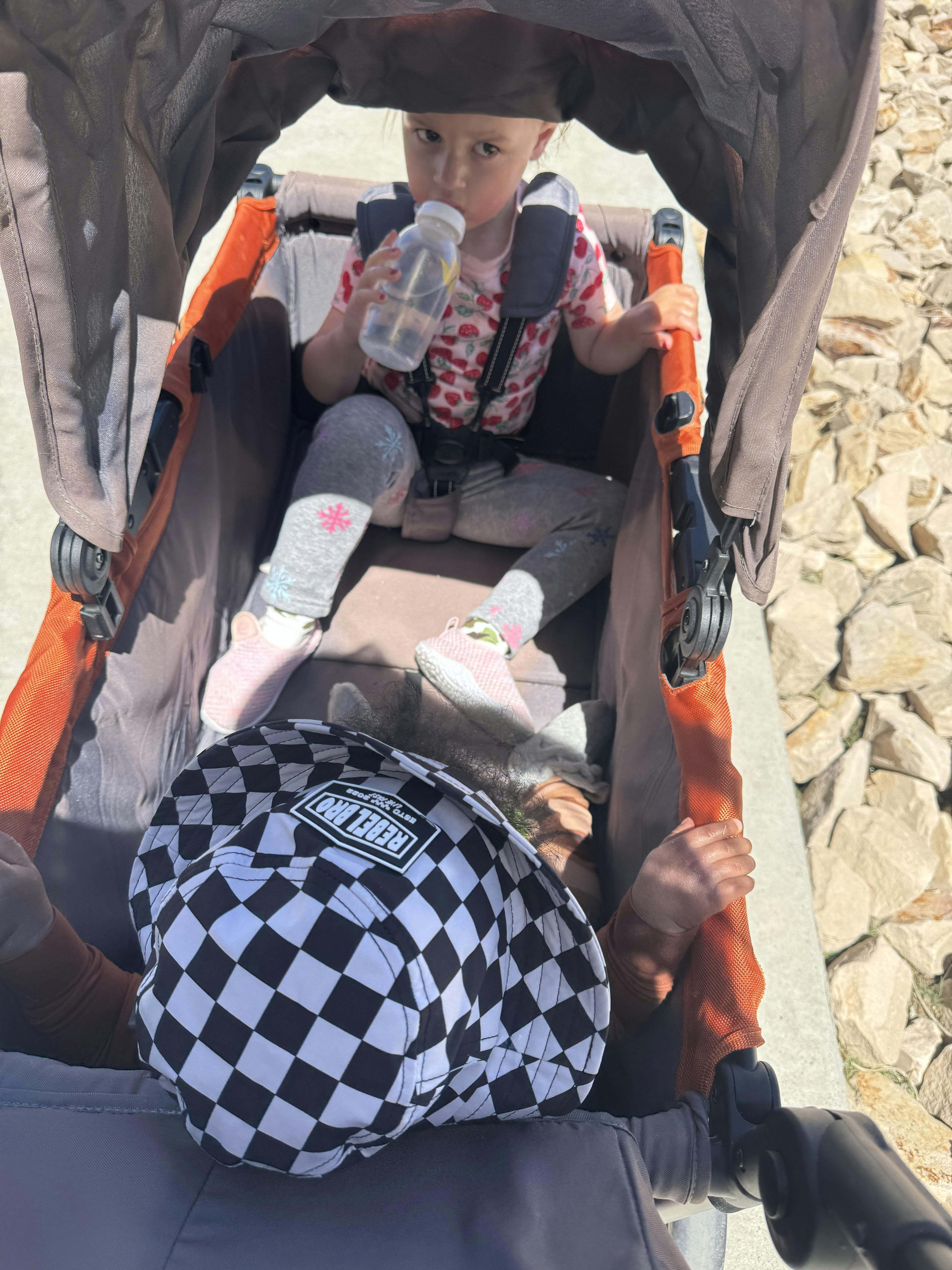 Child drinking from a bottle in a stroller with another child wearing a checkered hat