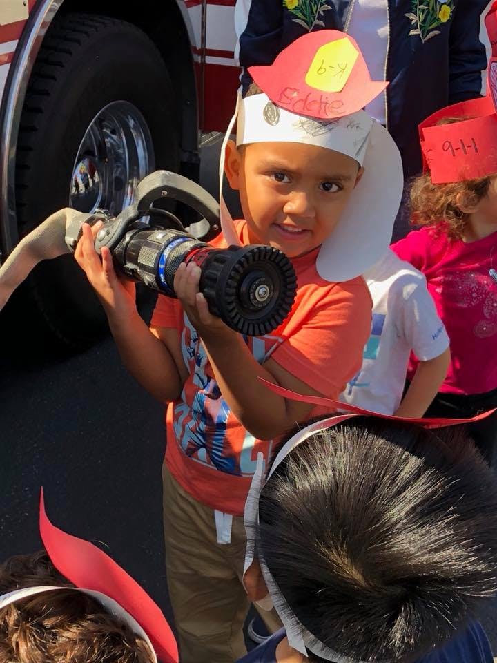 Child in a paper firefighter hat holding a fire hose nozzle