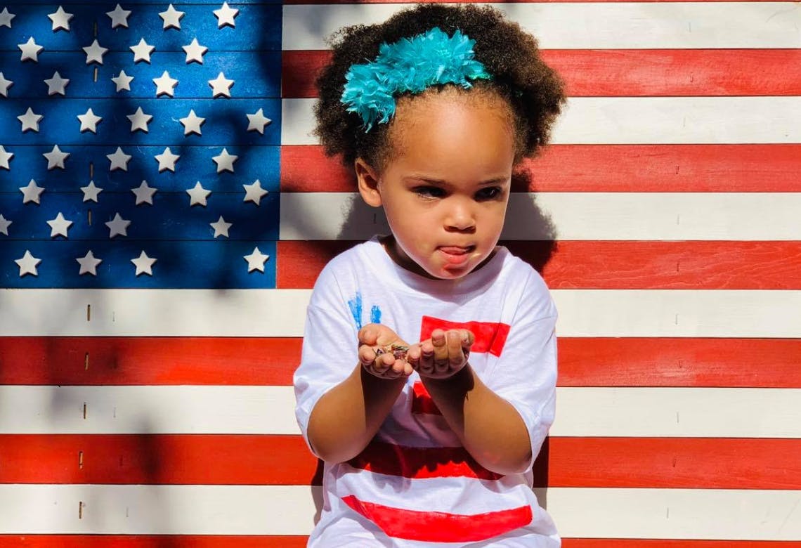 Child with blue headband holding hands out in front of an American flag