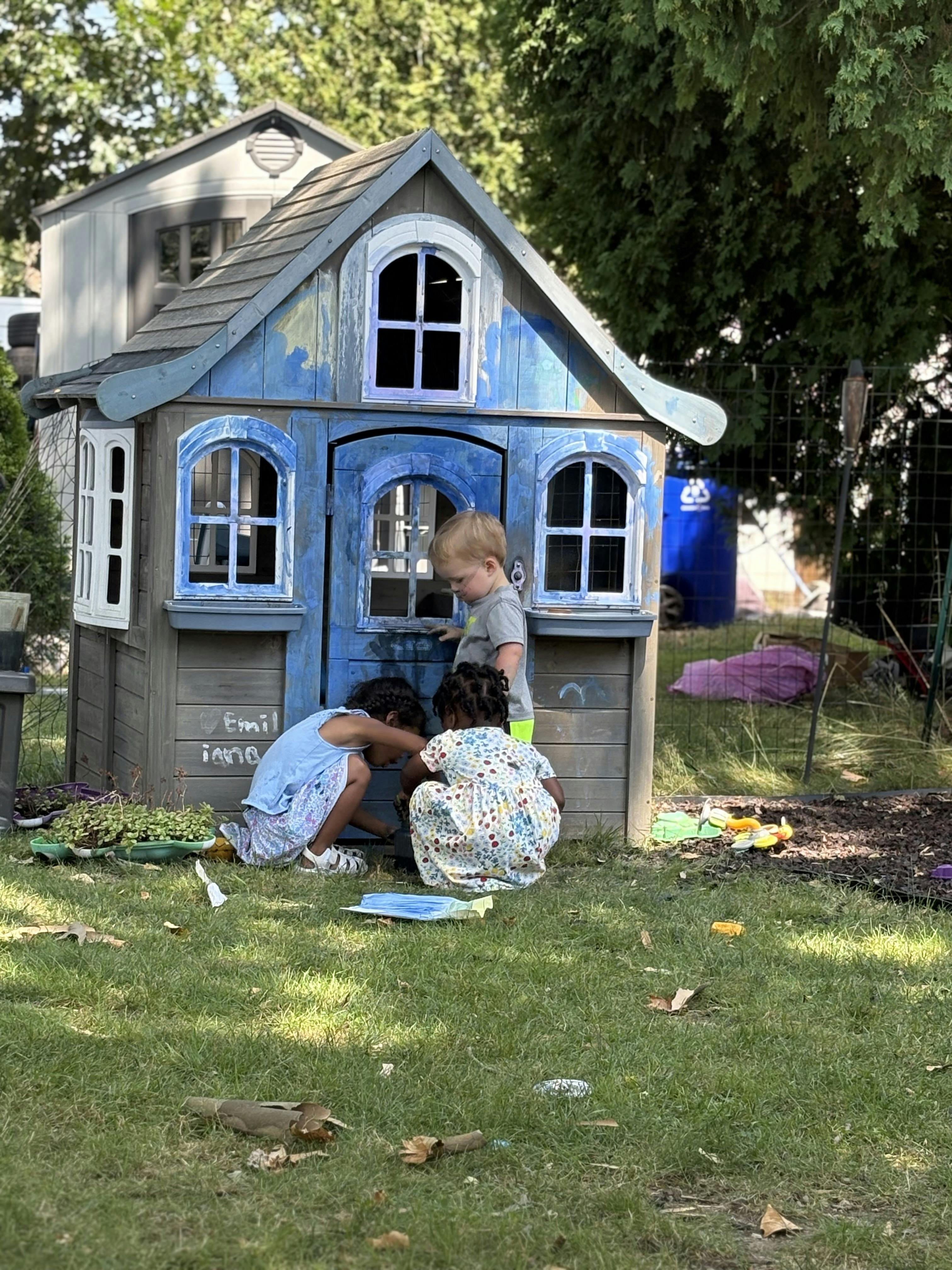 Three children playing together in a blue wooden playhouse in a backyard