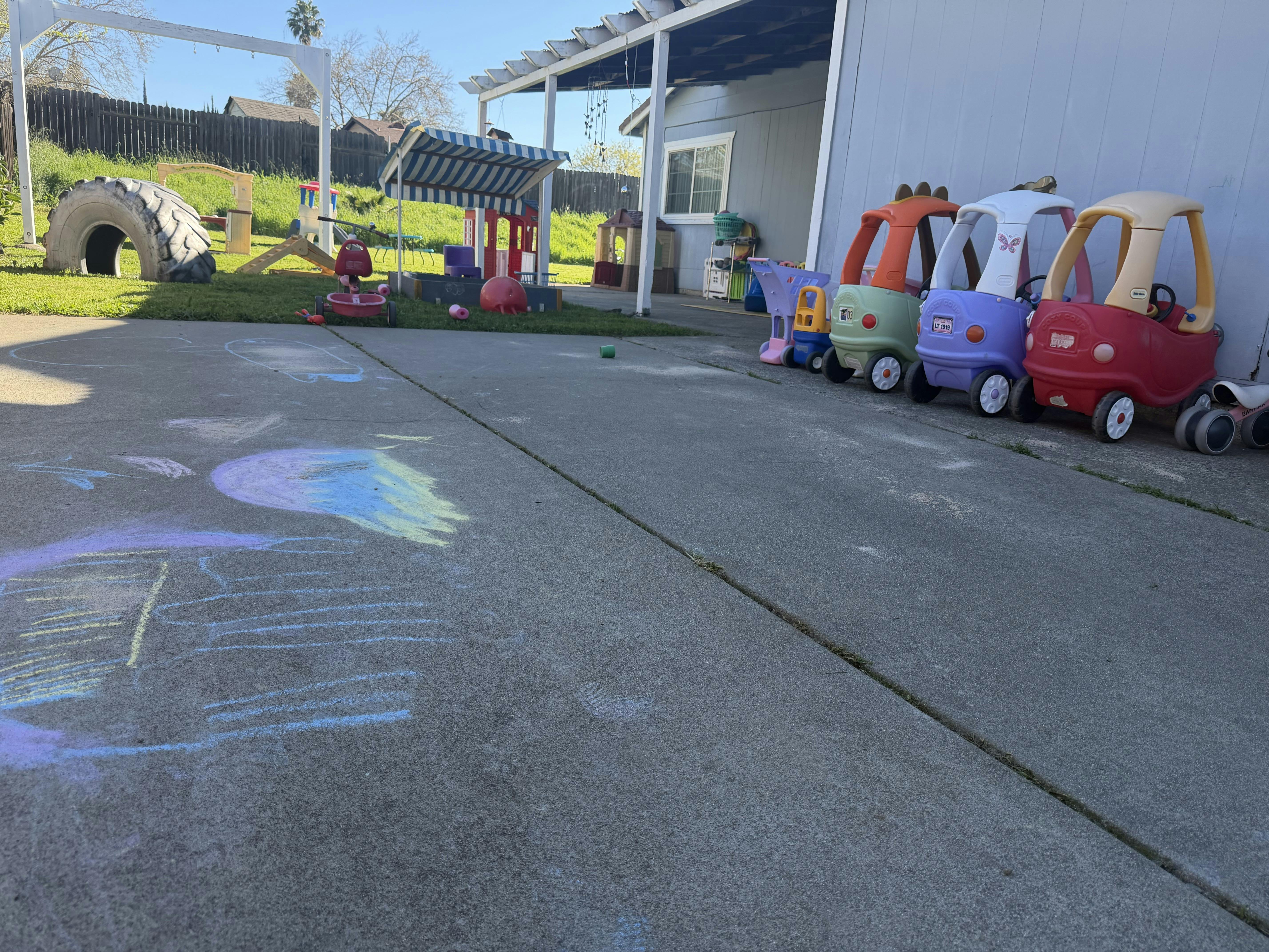 Children's ride-on cars next to a building, with playground equipment in a yard