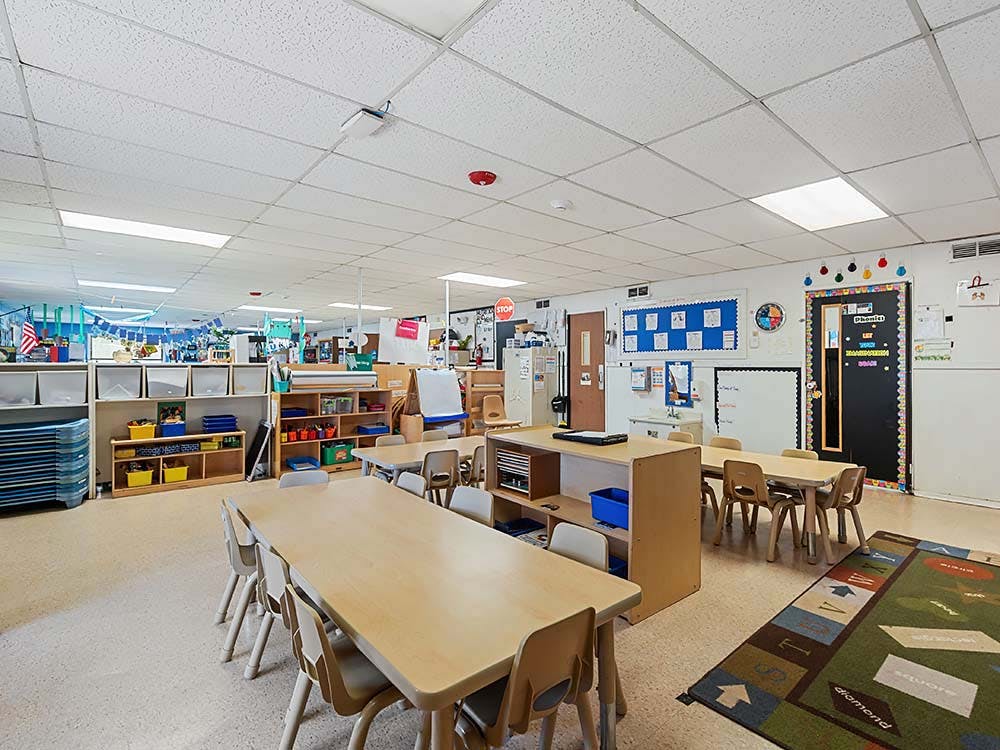 Empty classroom with small tables and chairs, storage shelves, and learning rug