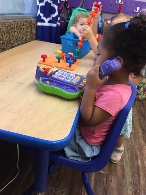 Child playing with a toy phone at a table, another child playing in the background