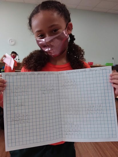 Child in a mask holding up a sheet of graph paper with writing