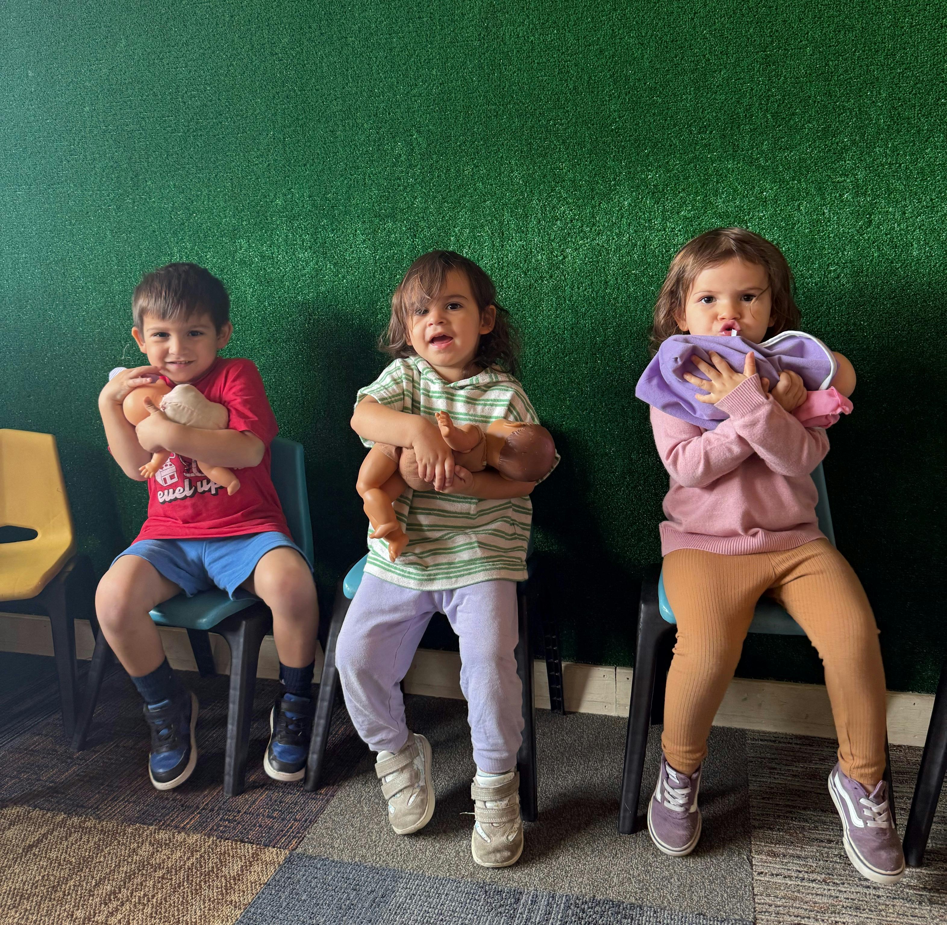 Three children sitting on chairs holding dolls