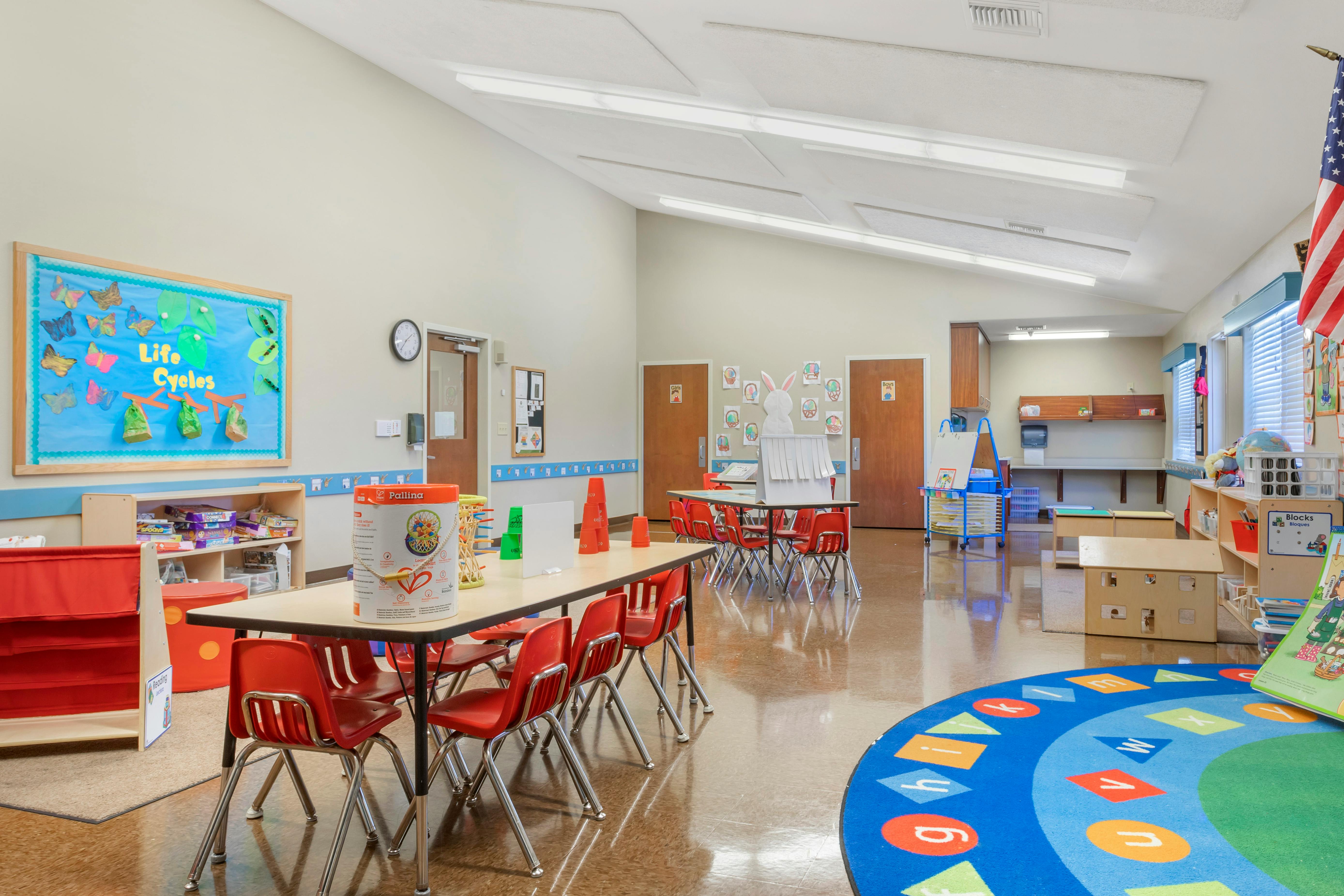 Classroom with child-sized tables and chairs, a colorful rug, and bulletin boards