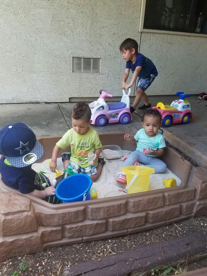 Three children playing in a sandbox with toys, one child pushing a toy car