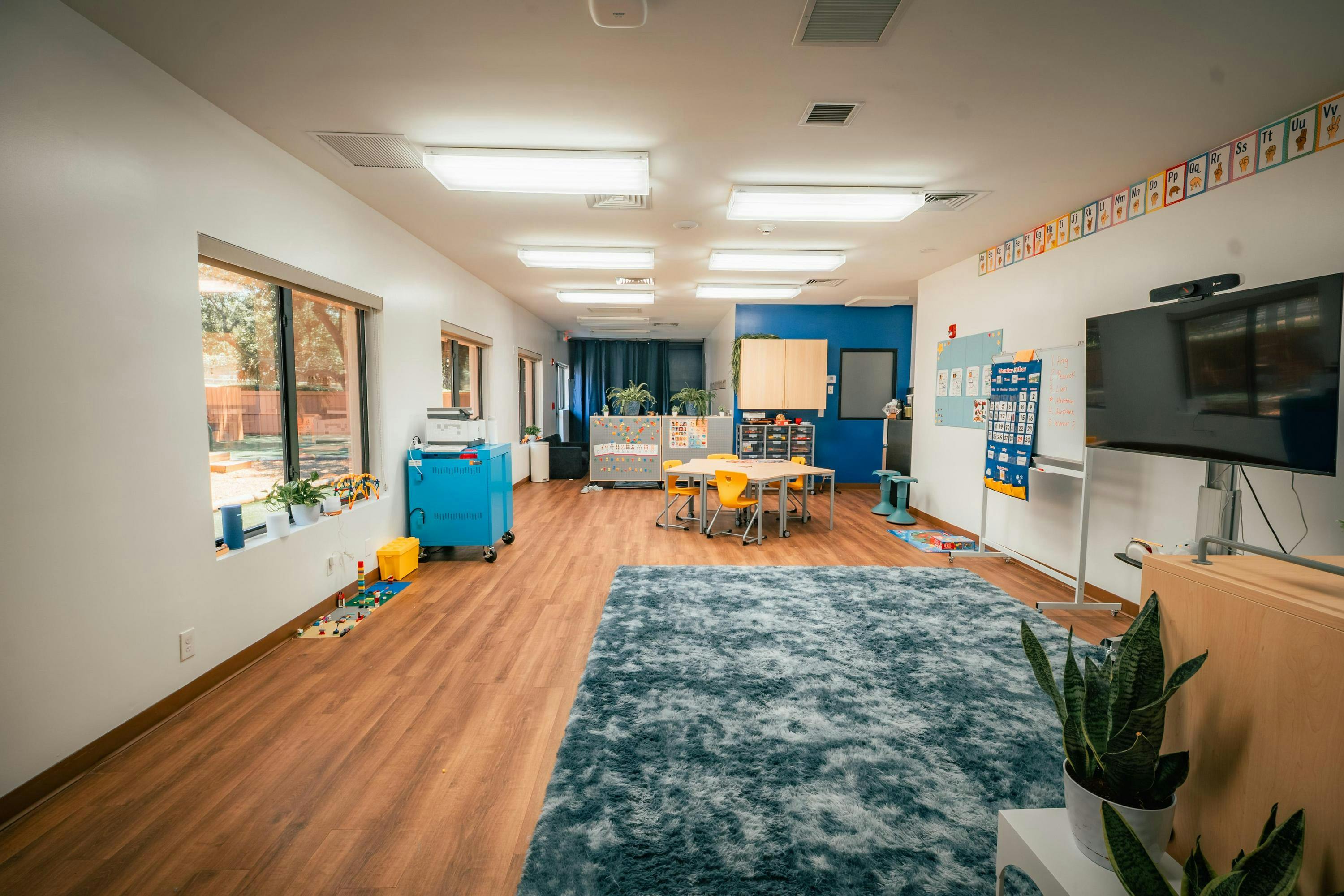 Classroom with large windows, bright lights, blue rug, and tables with yellow chairs