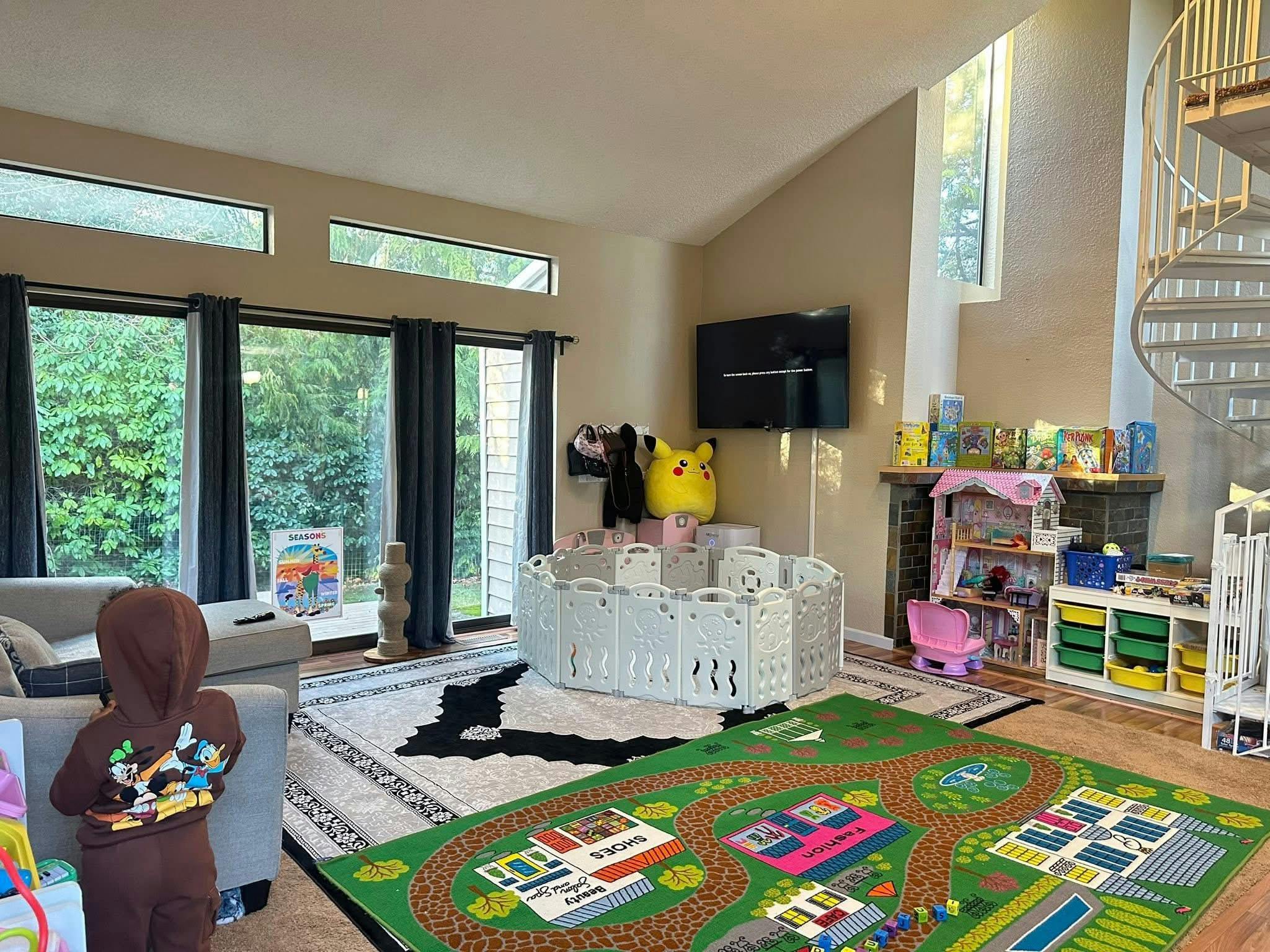 Child standing in a living room with a playpen, toy house, and rug with roads