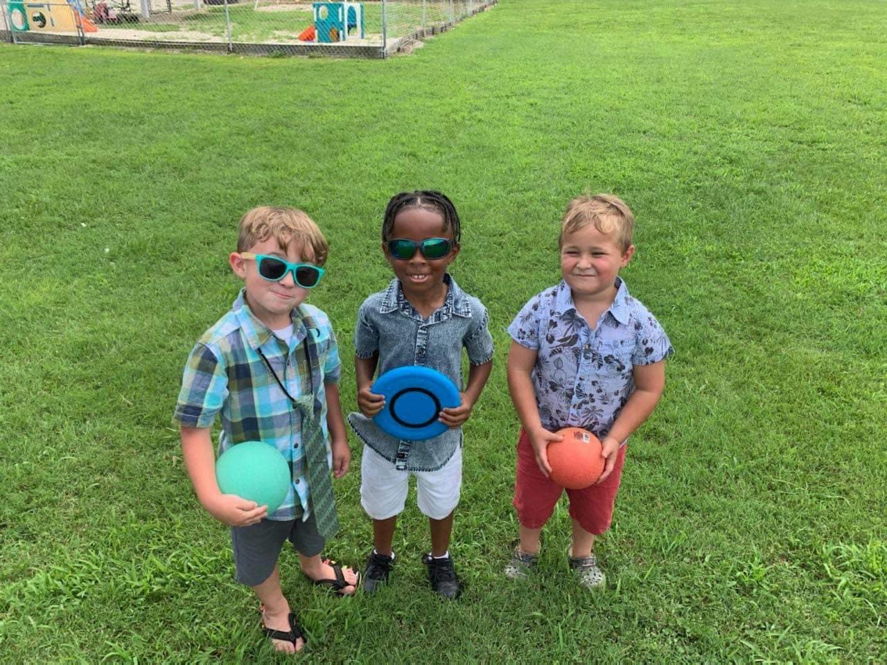 Three children in sunglasses holding balls and a frisbee on grass