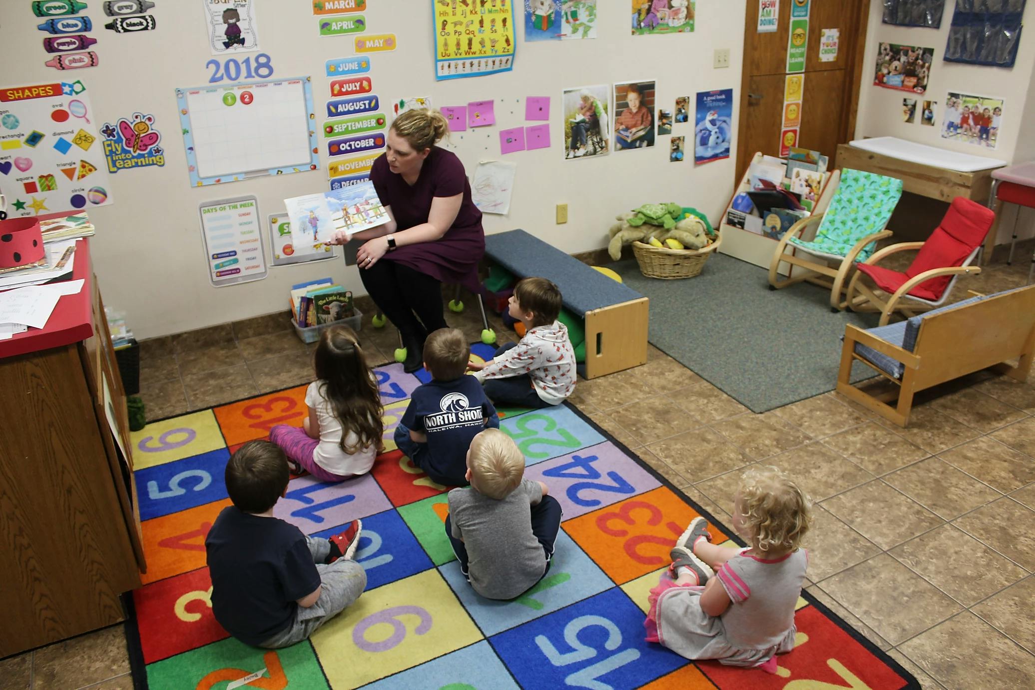 Person reading a book to children sitting on a colorful rug in a classroom