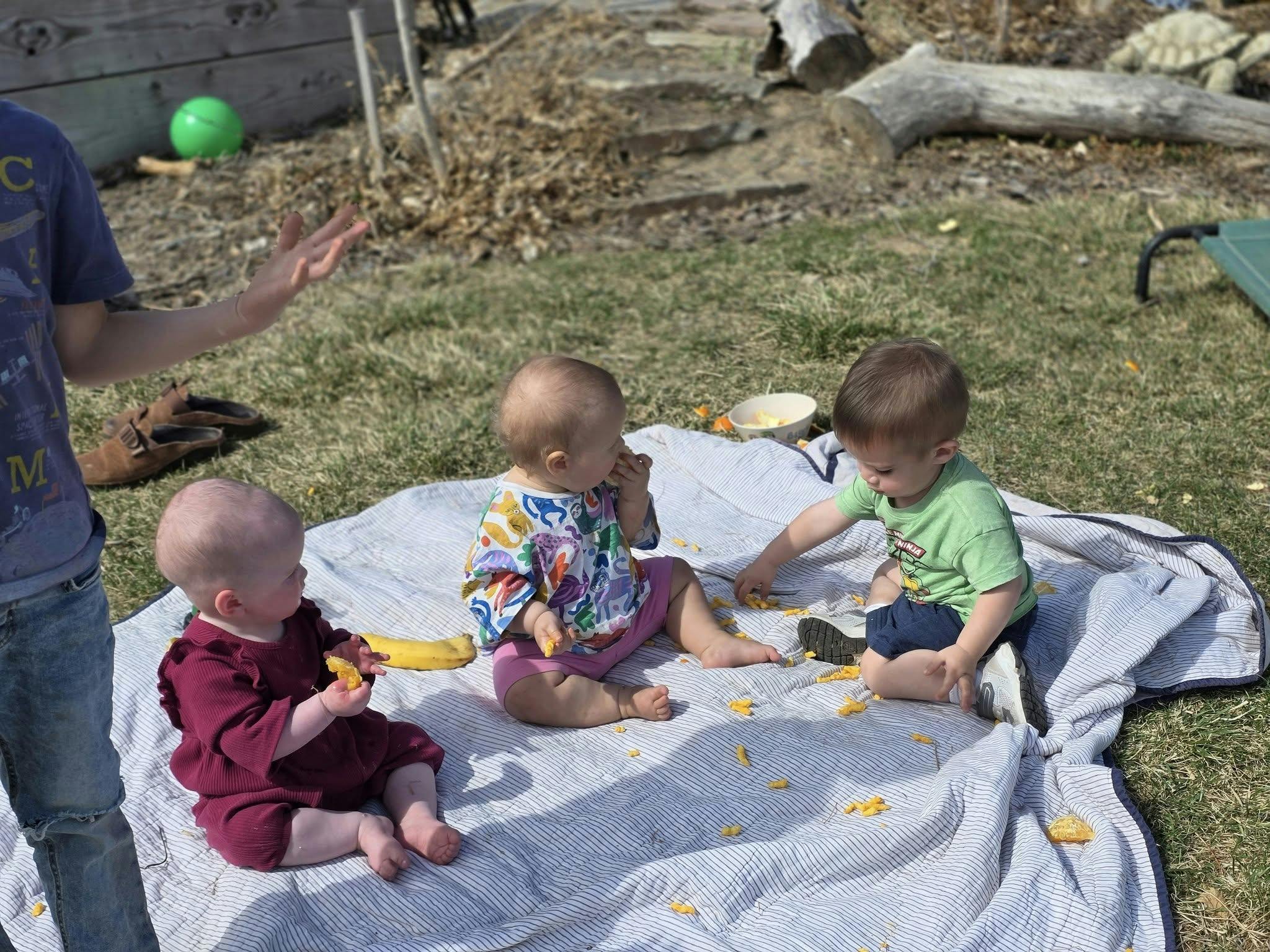 Three children sitting on a blanket outdoors, one person standing nearby