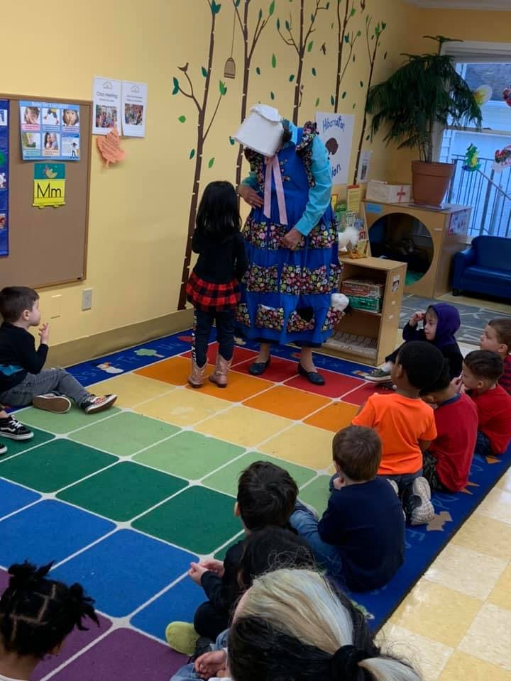 Adult in costume interacting with a child in a classroom with other children sitting on a rug