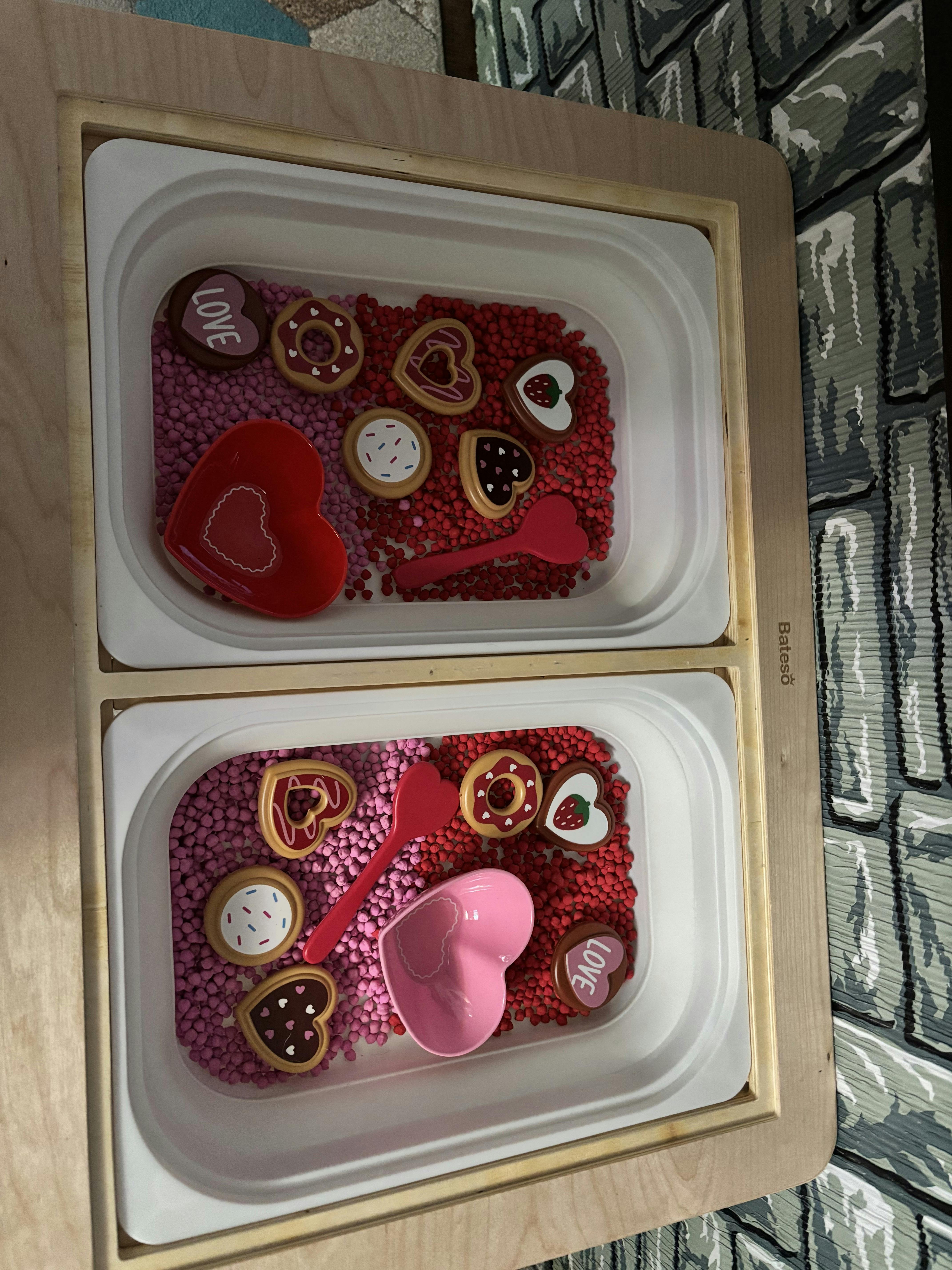 A sensory bin with red and pink beads, heart-shaped bowls, spoons, and cookies