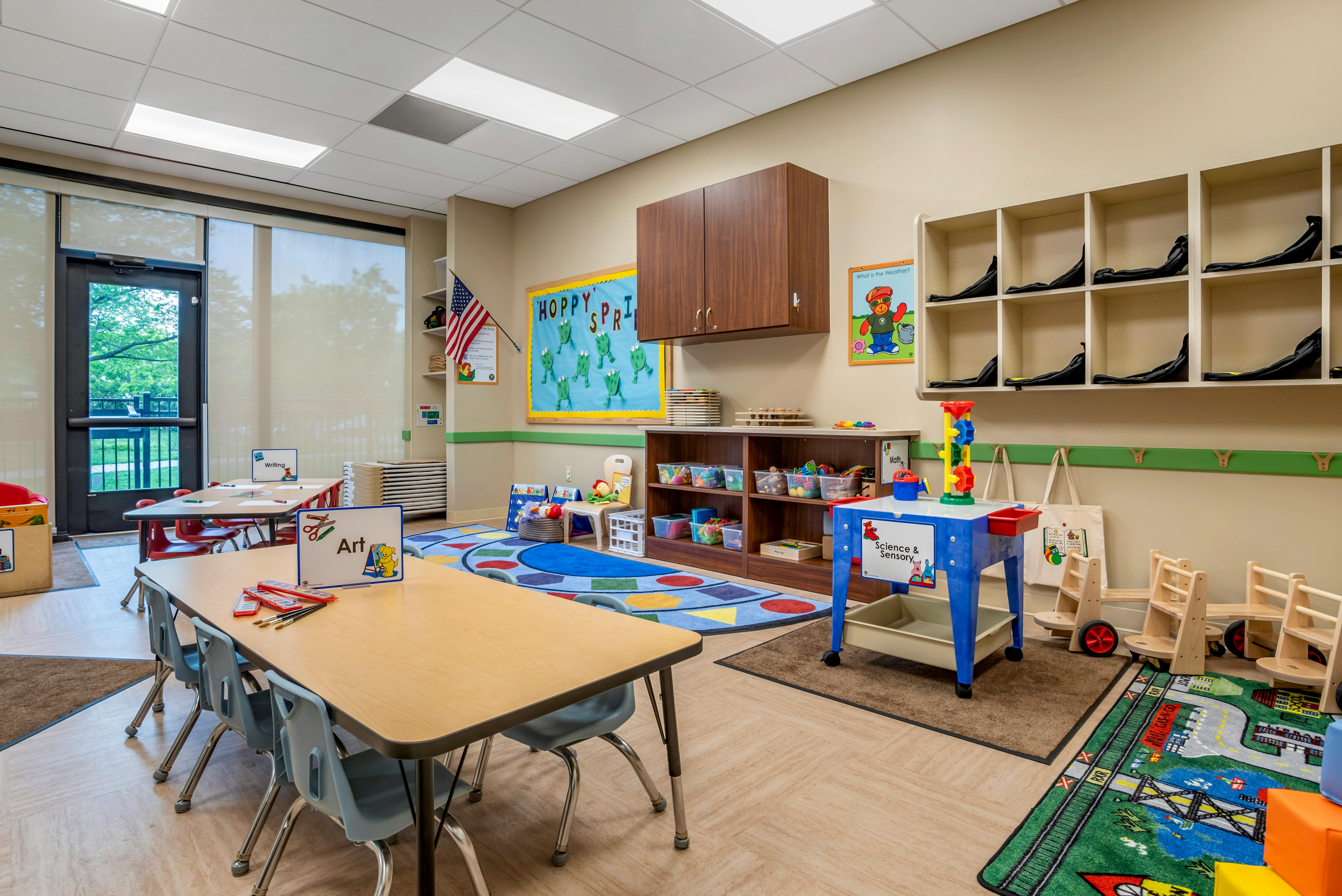 An empty classroom with tables, chairs, cubbies, and various learning stations