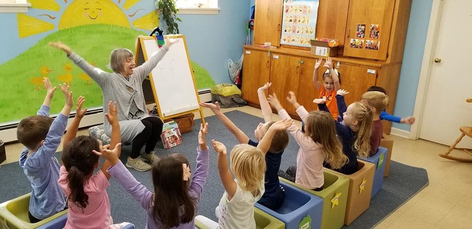 Person leading children in an activity with arms raised in a classroom