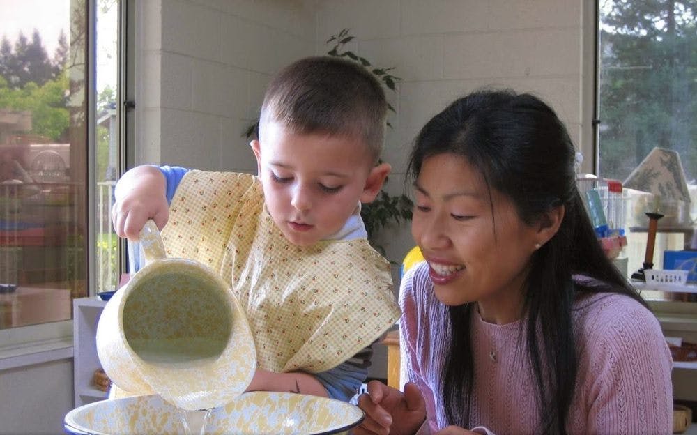 Child pouring from a pitcher into a bowl with a person watching