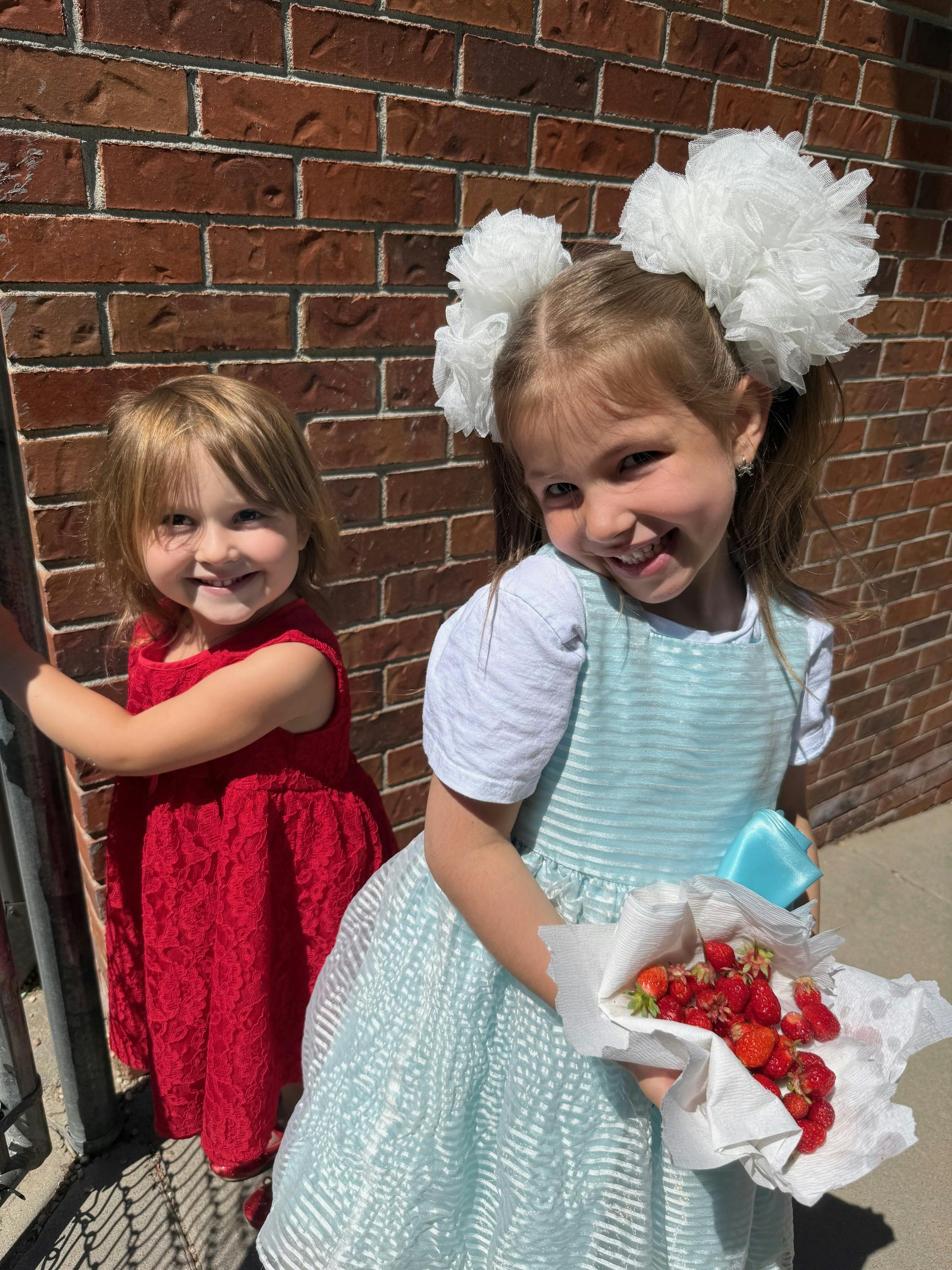 Two children standing by a brick wall with one holding a handful of strawberries