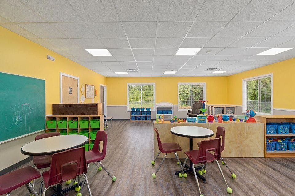 Empty classroom with small tables and chairs, yellow walls, and a green chalkboard